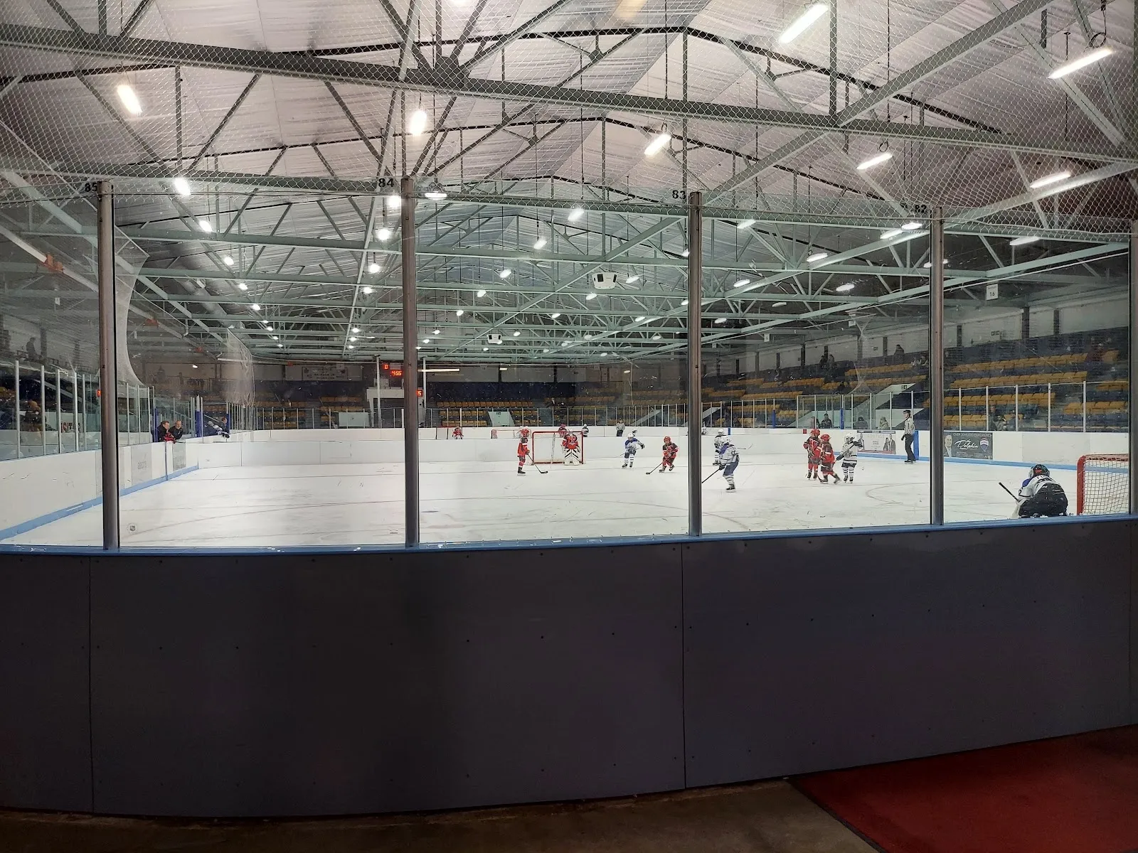 Jugadores con chamarras verdes alineados a lo largo de las vallas cercanas de la pista, con armaduras de acero en el techo arriba y la pared de la alberca visible más allá del vidrio. — Centennial Community Centre (Markham)