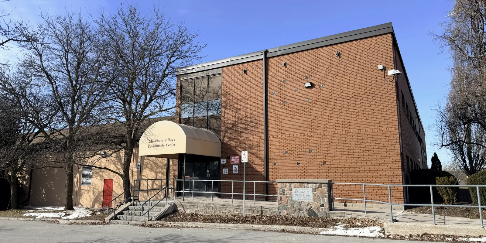 Front entrance of the brick Markham Village Community Centre building, with a tan awning reading "Markham Village Community Centre" over the glass doors and a stone "Home of Markham Fair" marker beside the steps. — Markham Village Arena
