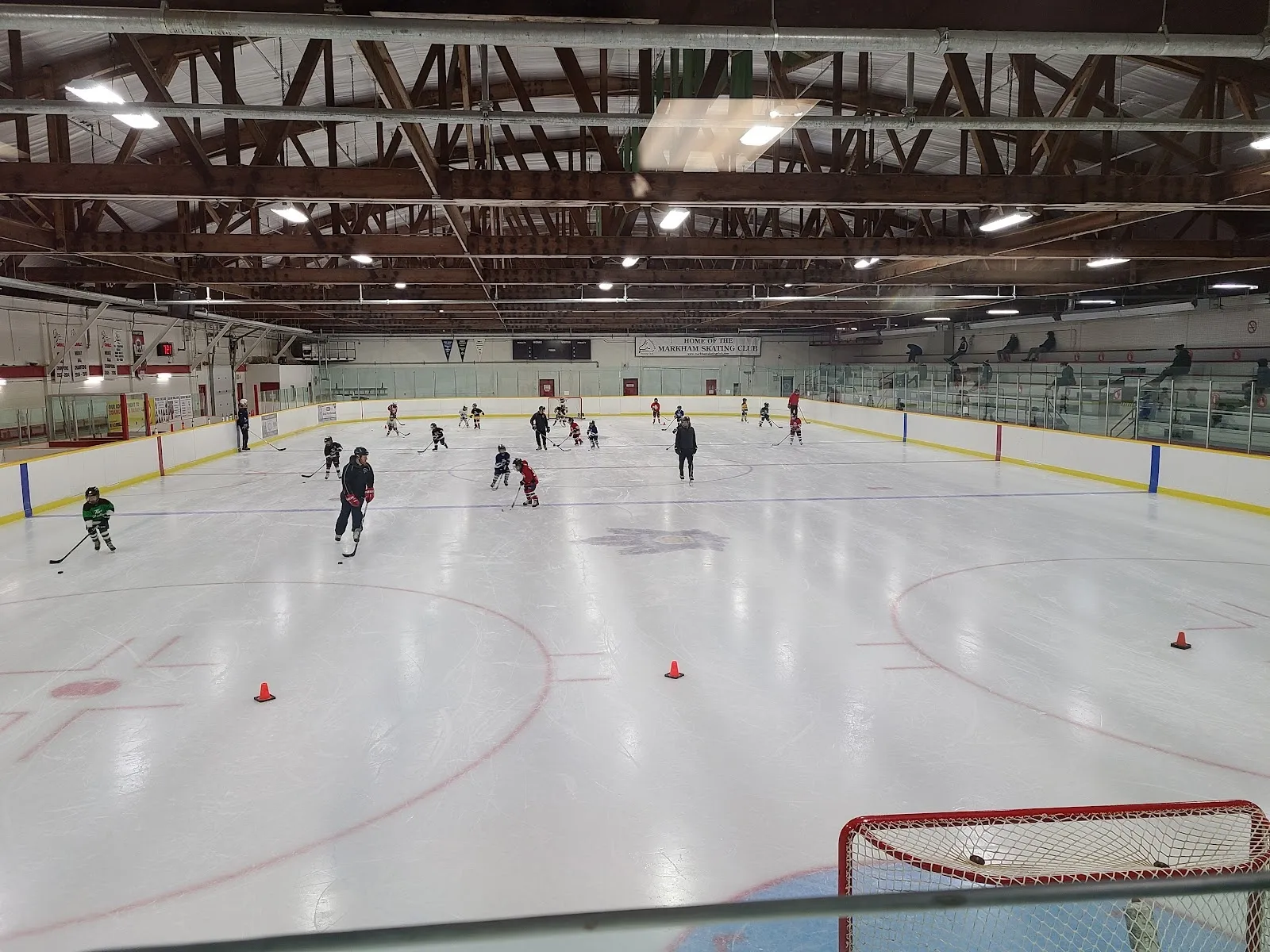 A public skating session spread across the sheet, cones and a goal in the foreground and a timber-trussed roof with banners along the far wall. — Markham Village Arena