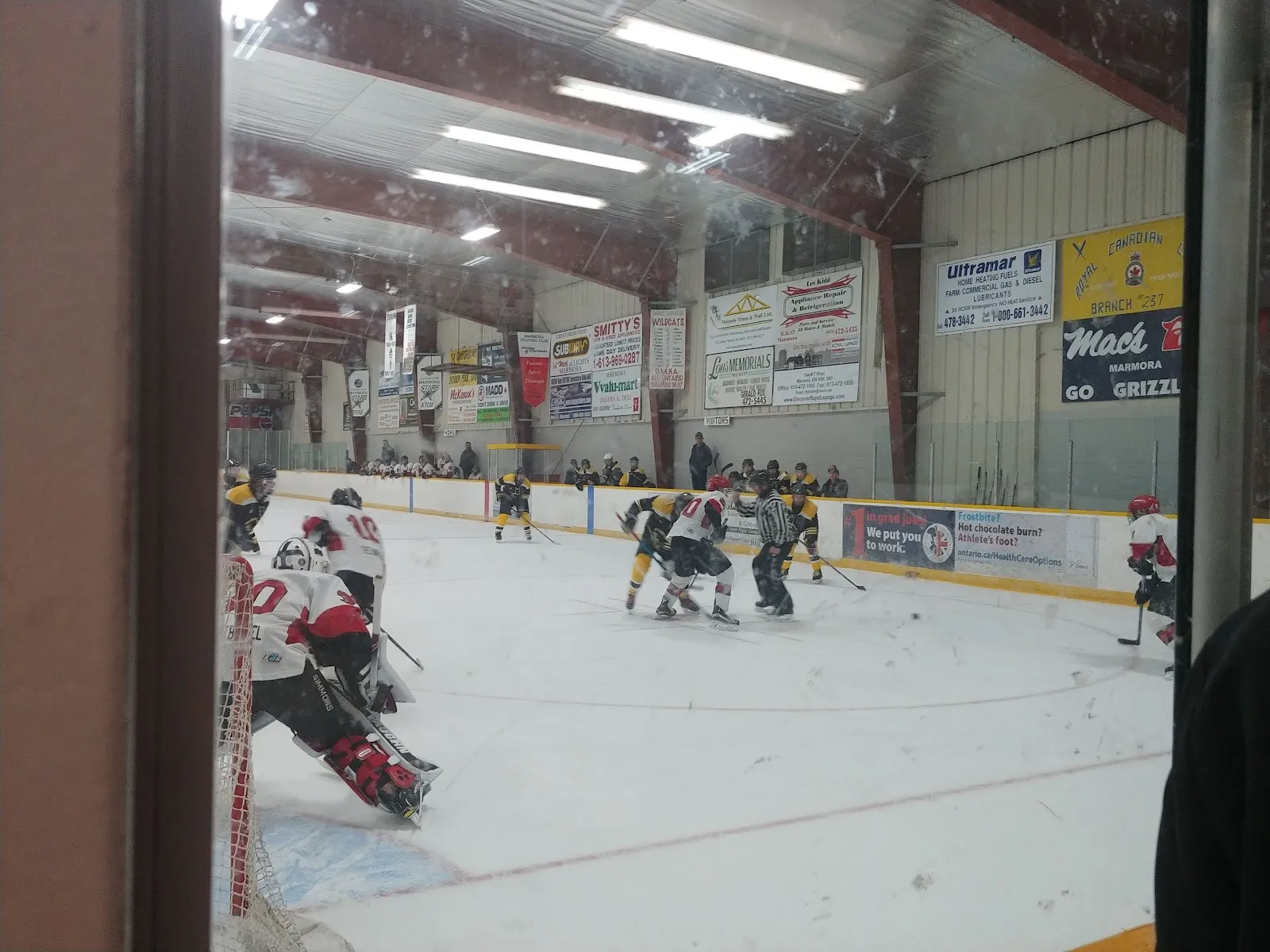 A minor hockey game in play at Marmora arena, seen through the glass at the end of the sheet, with sponsor boards along the far wall under a steel roof. — Dr. Hamilton Crawford Memorial Arena (Marmora)