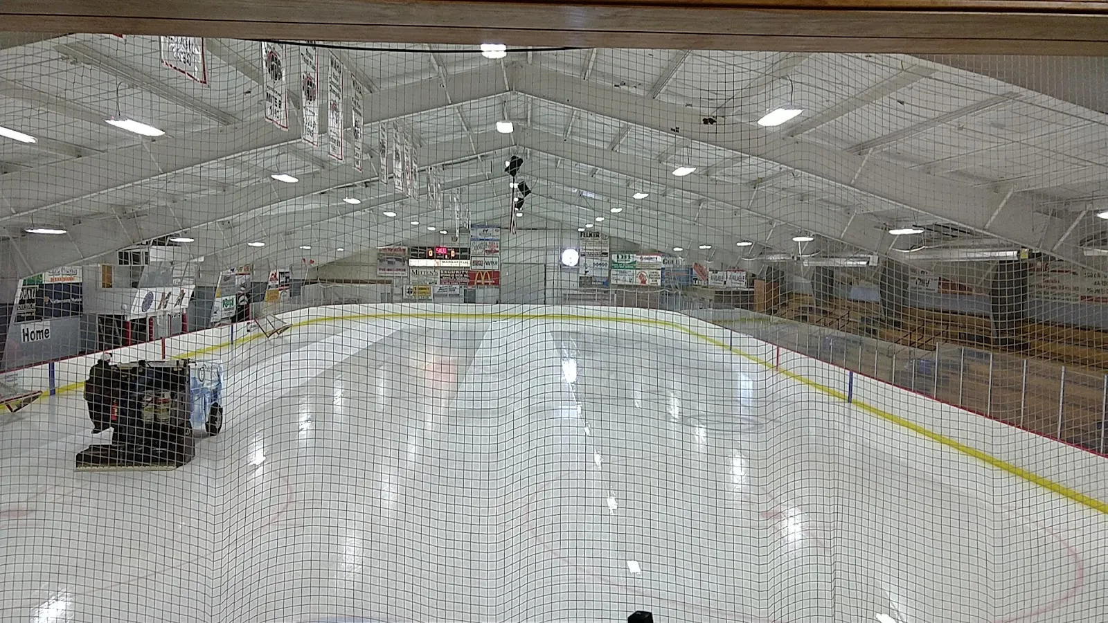 The ice sheet at Marshfield Area Ice Arena seen through the end netting, with the ice resurfacer and the roof trusses; no people — Marshfield Area Ice Arena