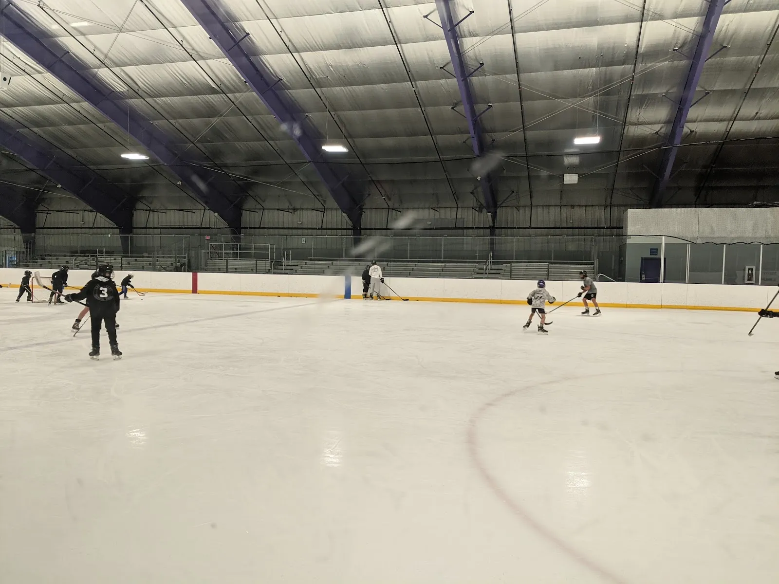 A wide view of the ice from one end with a few skaters and players spread across the sheet under purple steel trusses and a high white roof. — Martha's Vineyard Ice Arena