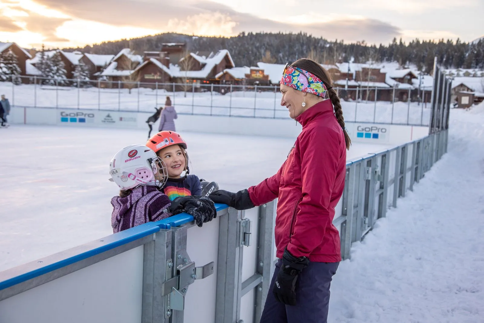 The outdoor rink in Big Sky Community Park at dusk, with young skaters at the boards and the snow-covered buildings of the town behind. — Marty Pavelich Ice Rink