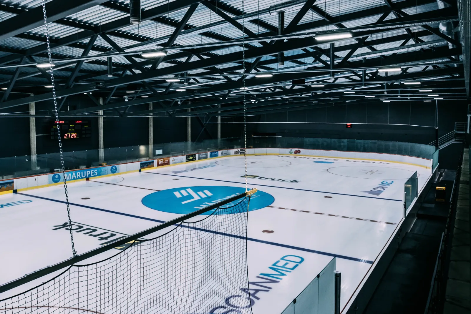 Mārupes Ledus halles tukšais ledus laukums, skatoties no tribīnēm, ar bortiem, centra logotipu un tērauda jumta kopnēm. – Mārupes Ledus Halle