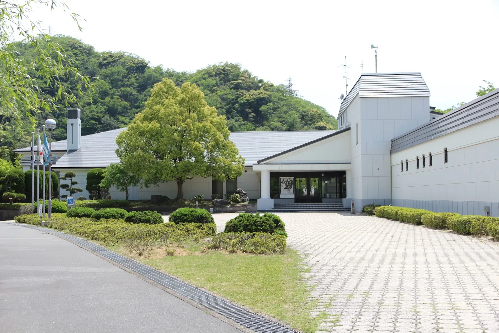 兵庫県立円山川公苑の入口の建物。舗装された前庭を備えた白い2階建ての施設で、背後には木々に覆われた丘が広がり、敷地内にはスケートリンクもあります。 — Maruyamagawa Kouen Skating Rink