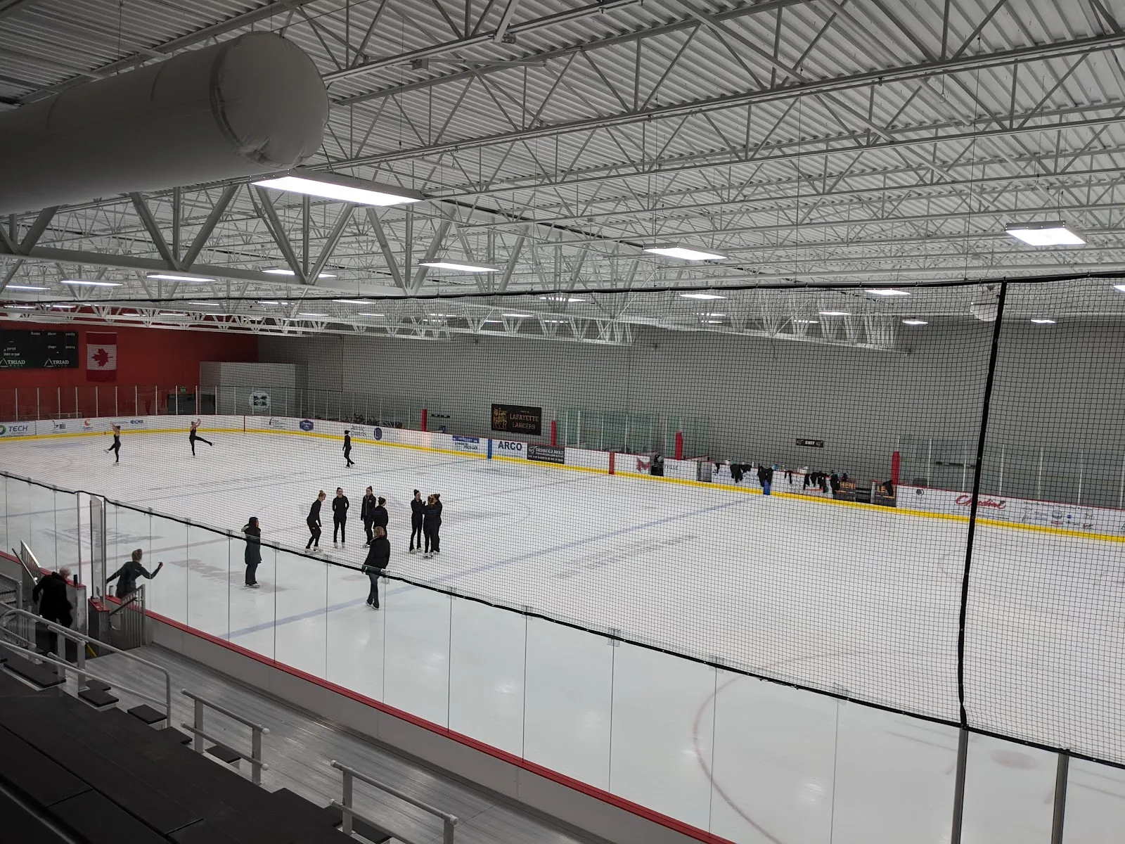 Figure skaters on the full ice sheet under the steel-truss roof, with a Lafayette Lancers banner and a Canadian flag hung above the boards — Maryville University Hockey Center