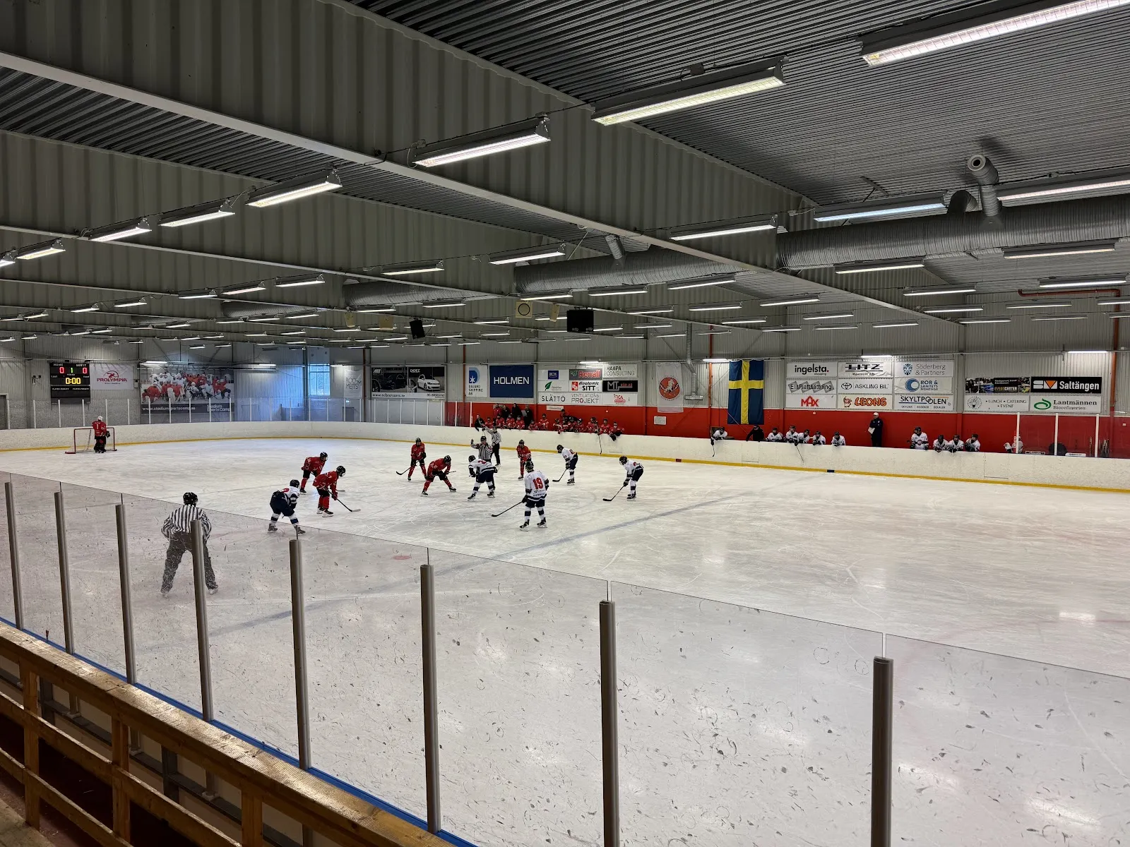 Vy över den inomhusbelägna isen med en hockeymatch igång och sargen och sponsorbanderollerna runt rinken. – Mässhallens islada Stallet