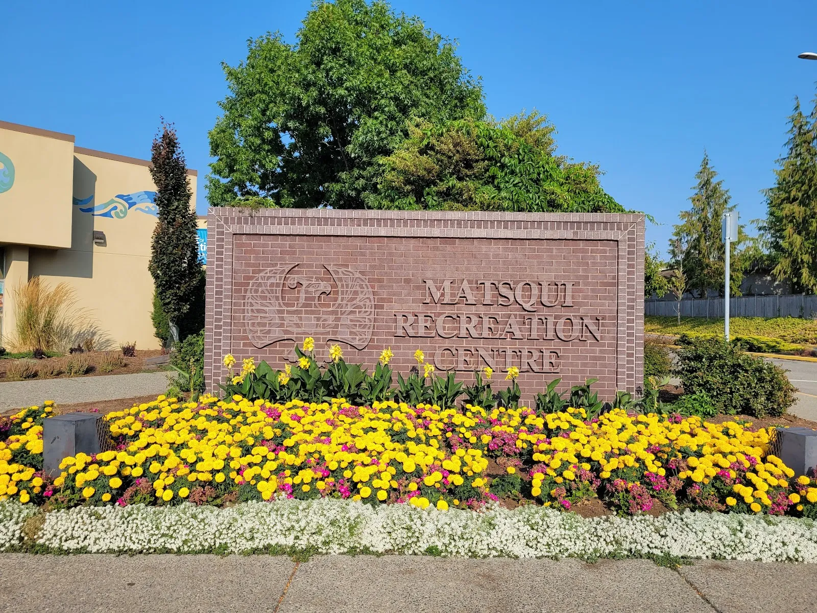 The brick Matsqui Recreation Centre entrance sign in a bed of yellow flowers in front of the building on a sunny day. — Matsqui Recreation Centre