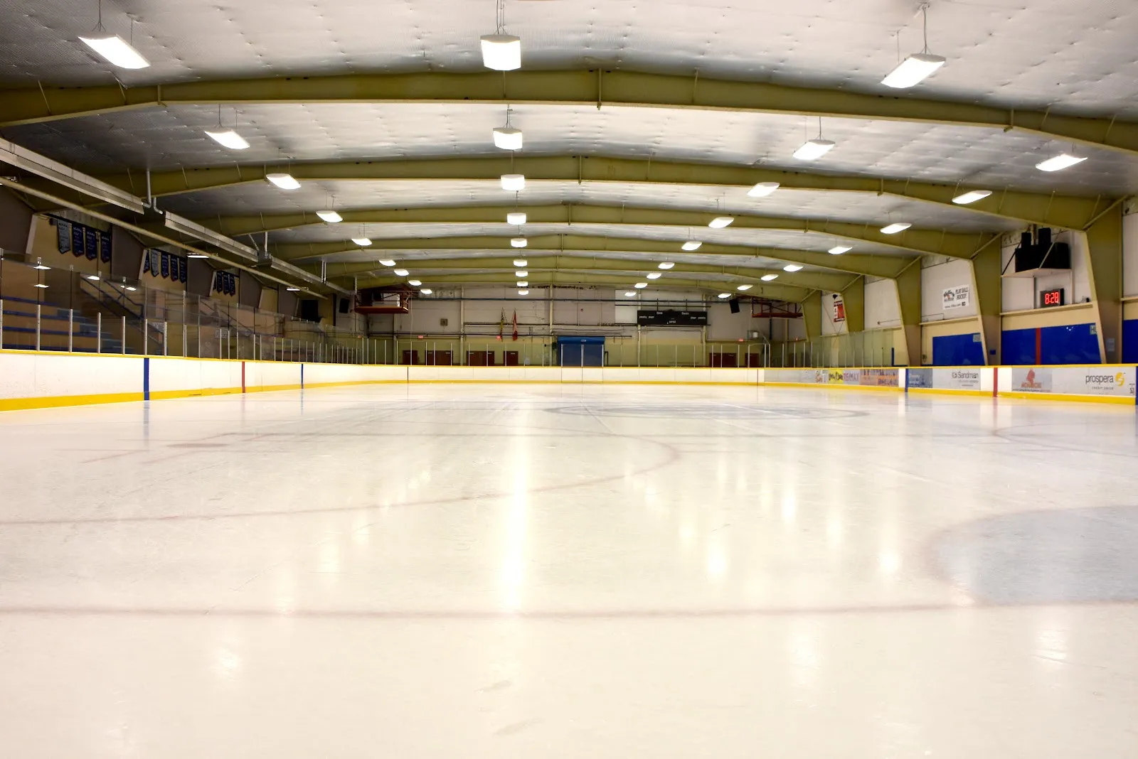 The empty ice sheet in the Matsqui Recreation Centre arena, with the boards, seating along the side wall and the arched roof trusses overhead. — Matsqui Recreation Centre