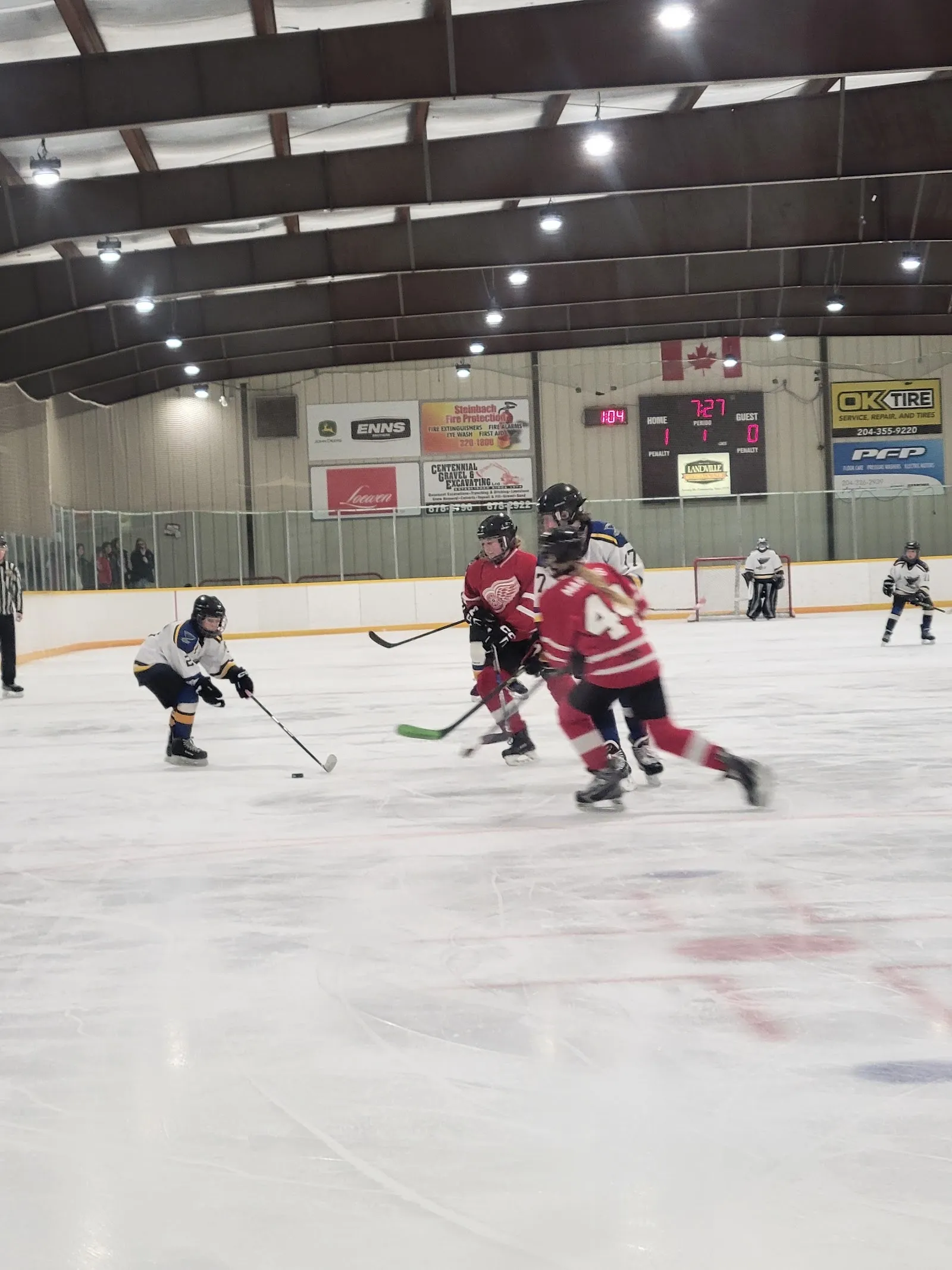 Youth hockey game in progress on the ice, with the scoreboard and sponsor boards along the far wall under the arena's dark roof beams. — Maurice Chaput Arena