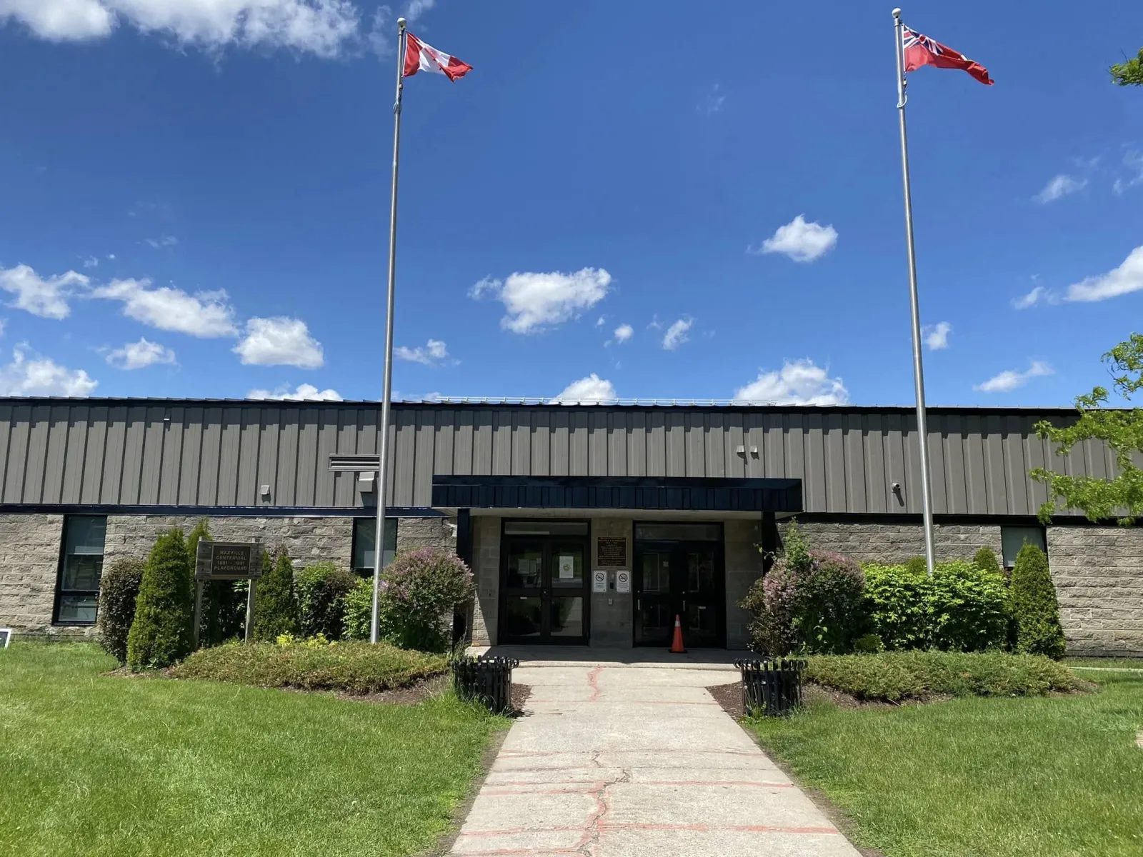 La entrada de un solo piso del Maxville District Sports Complex detrás de un césped y jardineras, con dos astas de bandera frente al techo oscuro de metal. — Maxville & District Sports Complex