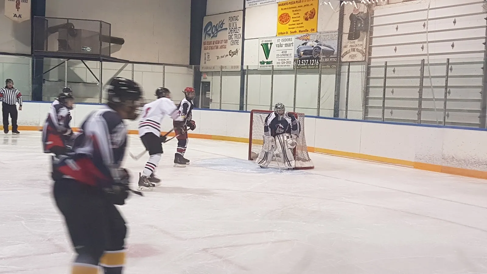 Un partido visto desde las gradas, con un portero y jugadores repartidos por la pista, una pared de banderines de Maxville y un barandal azul detrás. — Maxville & District Sports Complex