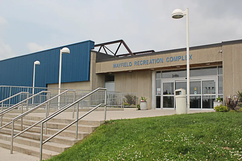 La entrada de Mayfield Recreation Complex con su nombre en la fachada de concreto, la pared metálica azul de la arena a la izquierda y escalones que suben desde el jardín. — Mayfield Recreation Complex