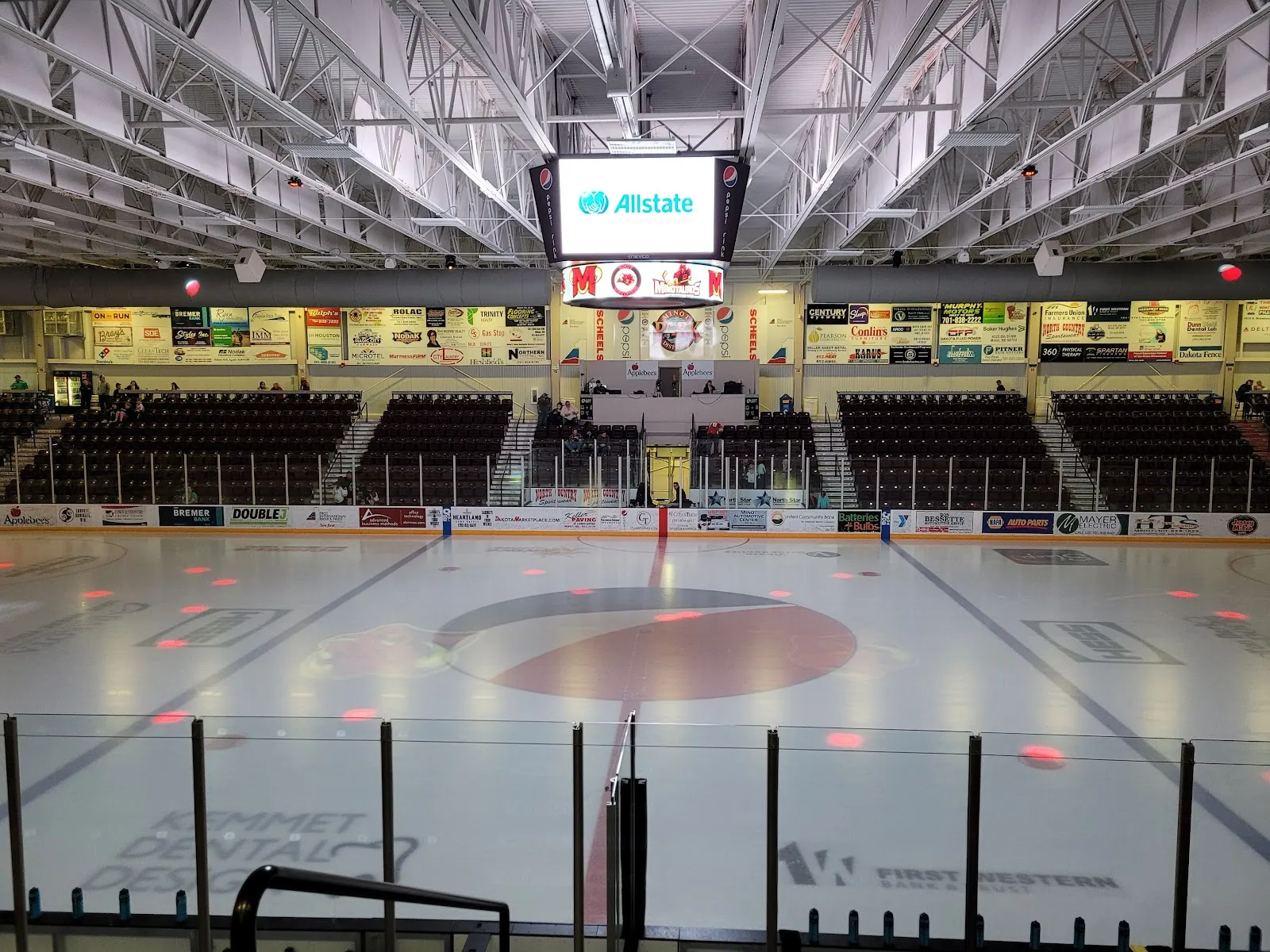 The empty Maysa Arena ice with centre-ice logo, scoreboard and lower bowl seating along both sides. — Maysa Arena