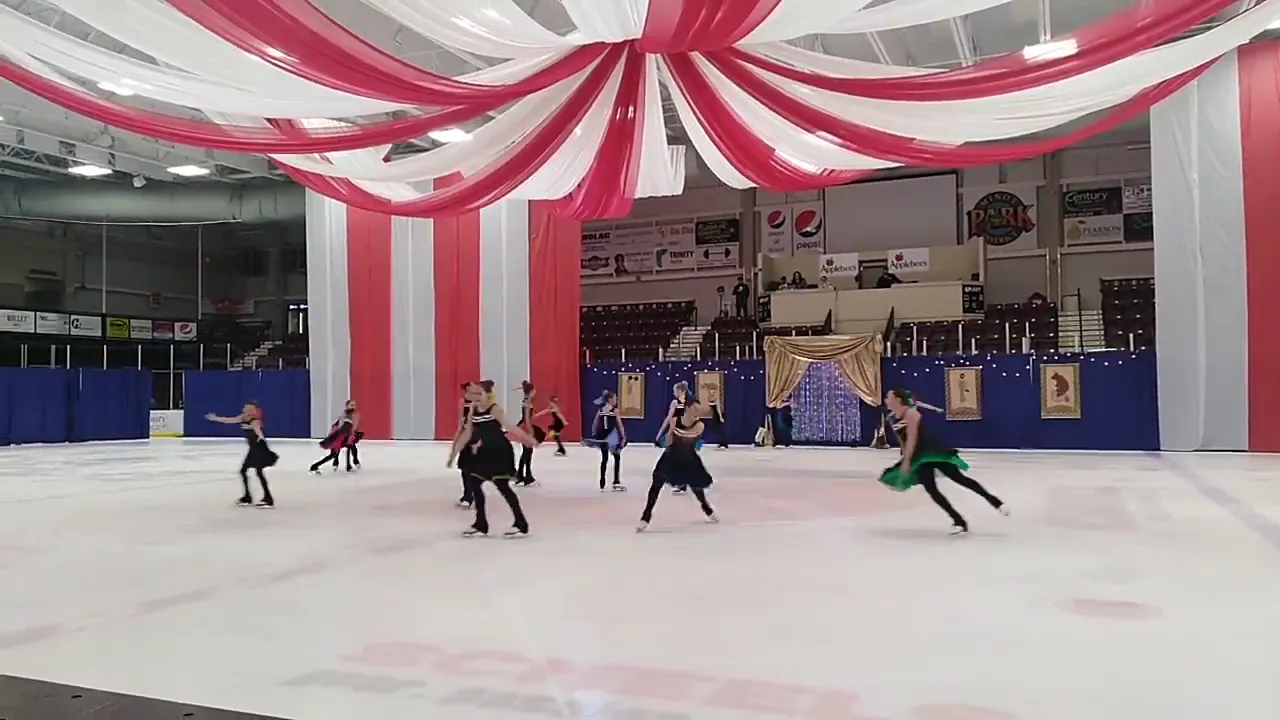 Skaters on the Maysa Arena ice under red and white striped banners, with the lower stands behind. — Maysa Arena