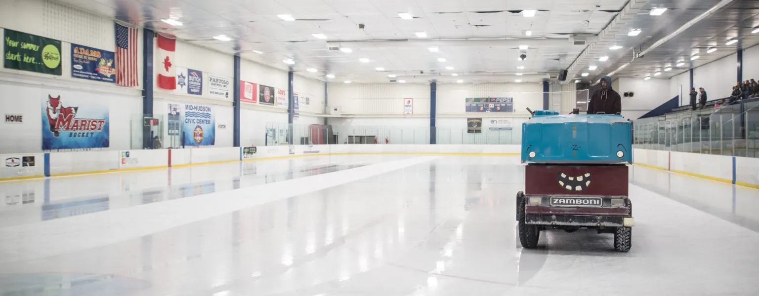 The empty rink under bright lights with an ice resurfacer crossing the fresh sheet, Marist banners and sponsor boards along the far wall. — McCann Ice Arena