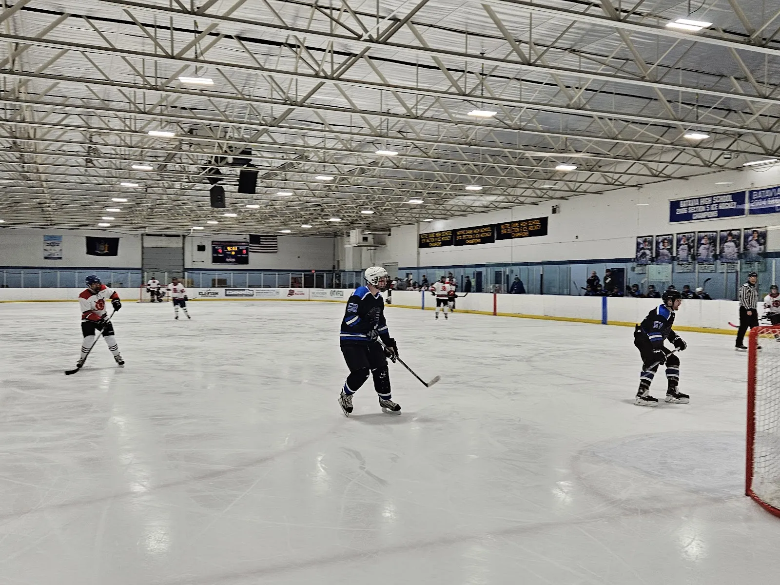 Un partido en el extremo lejano de una pista amplia, con jugadores repartidos por el hielo blanco bajo un largo techo de armaduras de acero con banderines de patrocinadores a lo largo de las vallas. — David M McCarthy Ice Arena