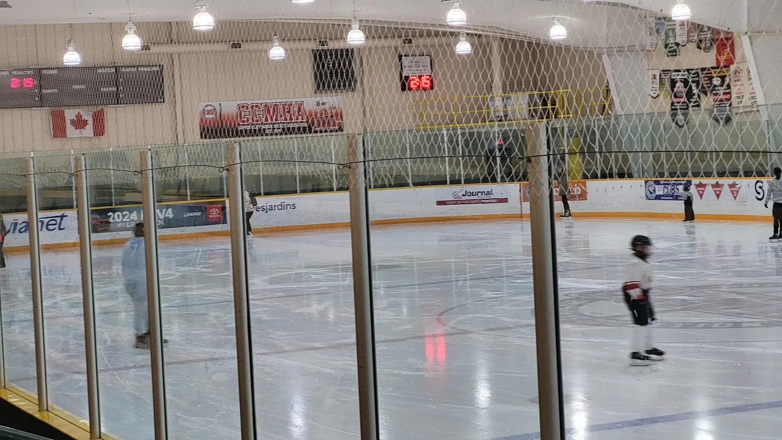 A game seen from a wooden bench along the rail, players spread across a wide sheet under a pale roof with Canadian flags on the far wall. — McClelland Community Centre and Arena (Copper Cliff)