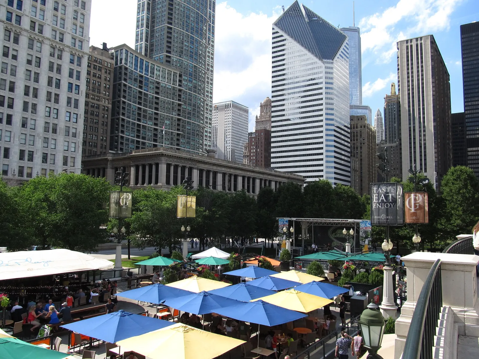 La plaza que ocupa la pista, vista desde el nivel de AT&T Plaza en verano, cuando la superficie de hielo es la terraza exterior del Park Grill con sombrillas, rodeada por los edificios de Michigan Avenue. — McCormick Tribune Ice Rink (Millennium Park)