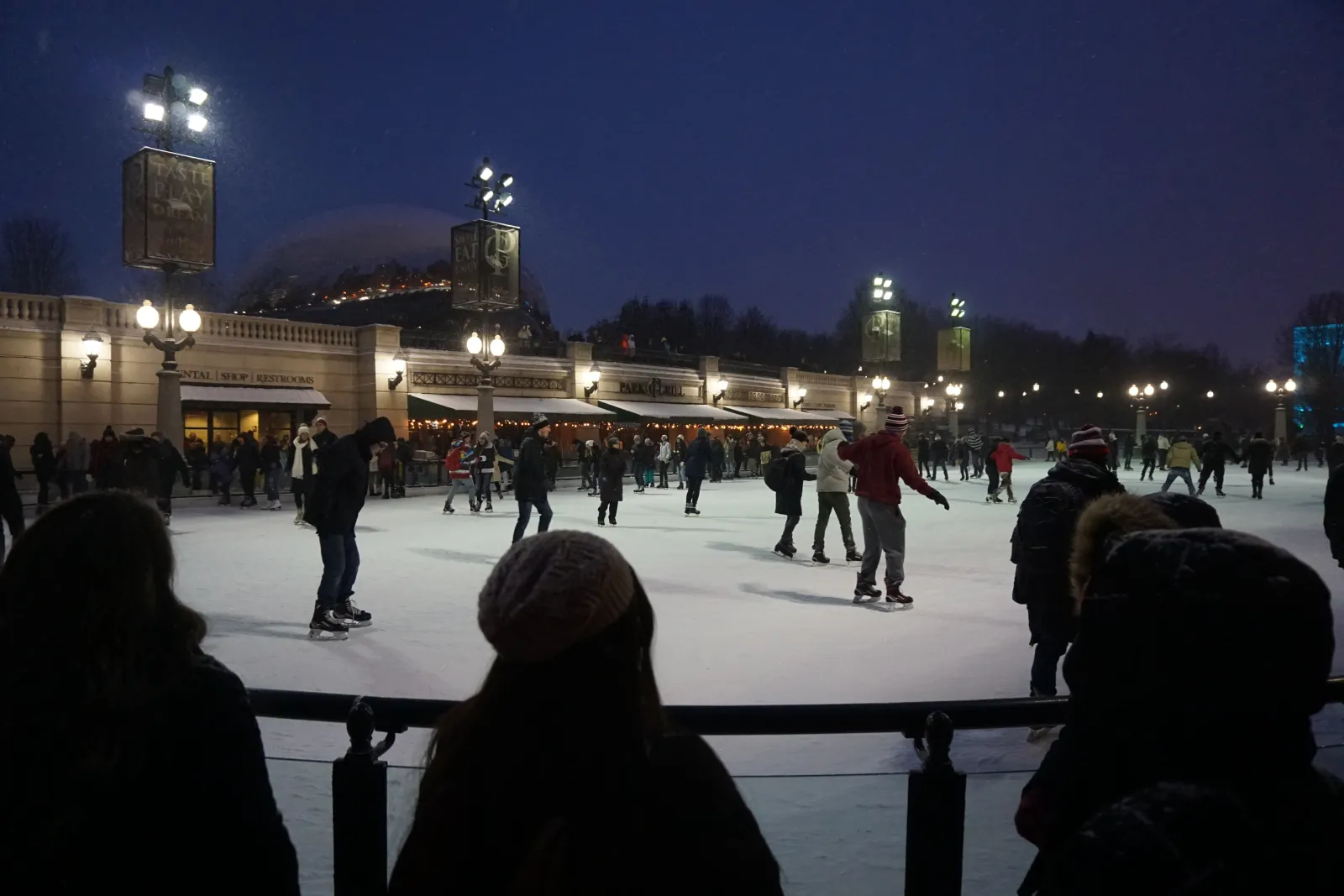 La pista de noche en diciembre de 2017, con patinadores sobre la superficie blanca bajo las lámparas, la columnata de la plaza detrás y espectadores en silueta junto a la baranda. — McCormick Tribune Ice Rink (Millennium Park)