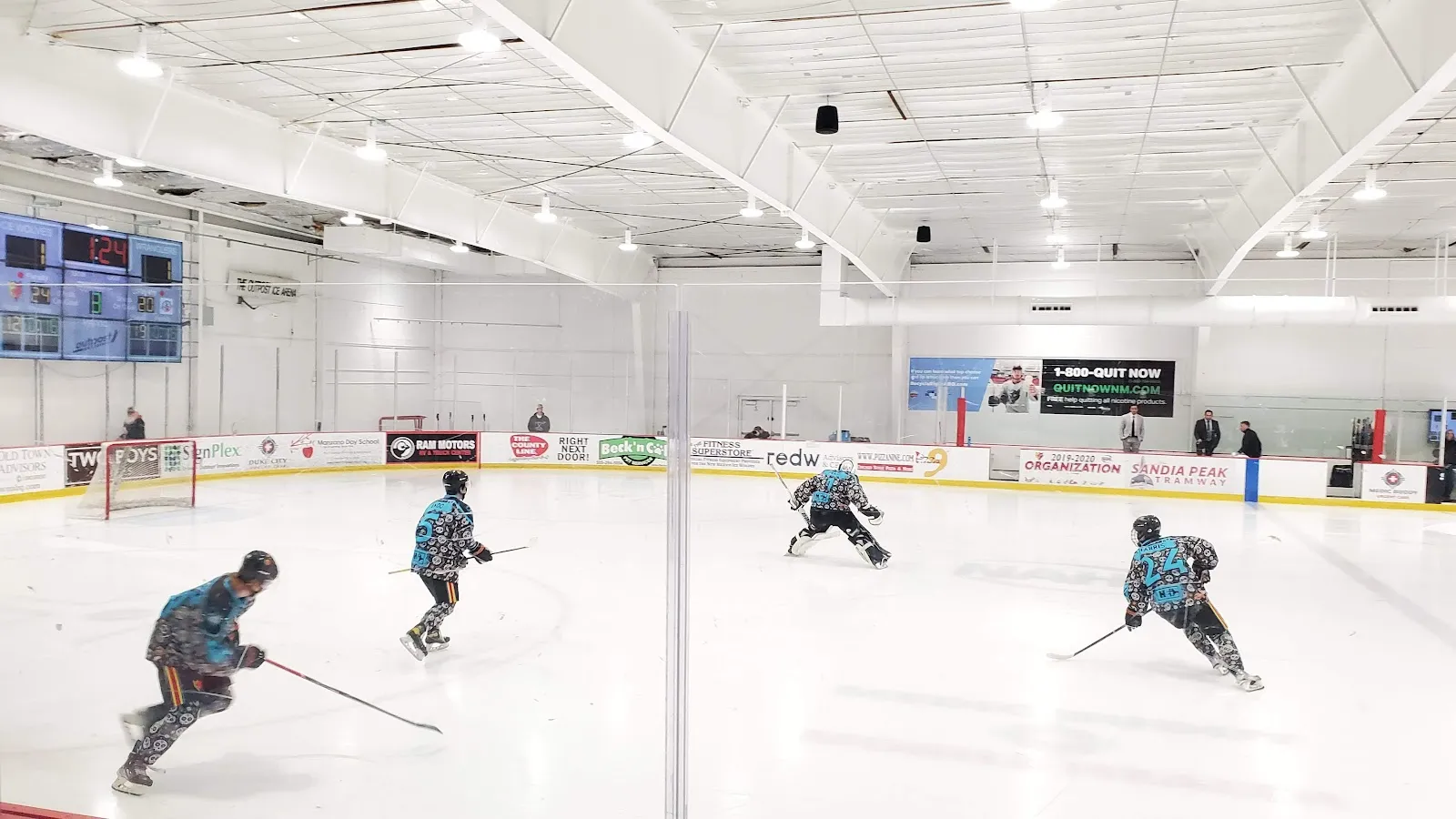 Skaters in teal jerseys playing on the full-size ice sheet, with the scoreboards and dasher boards behind them. — McDermott Athletic Center