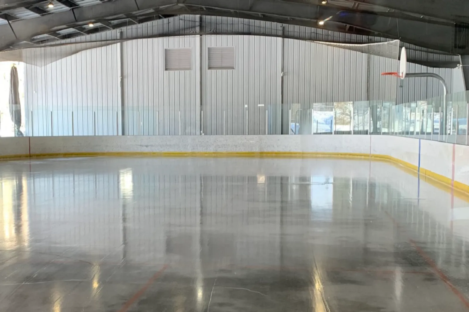 The ice sheet of the covered rink at the McDougall Recreation Centre, with white boards, glass panels and a basketball hoop under a metal roof. — McDougall Recreation Centre Outdoor Rink
