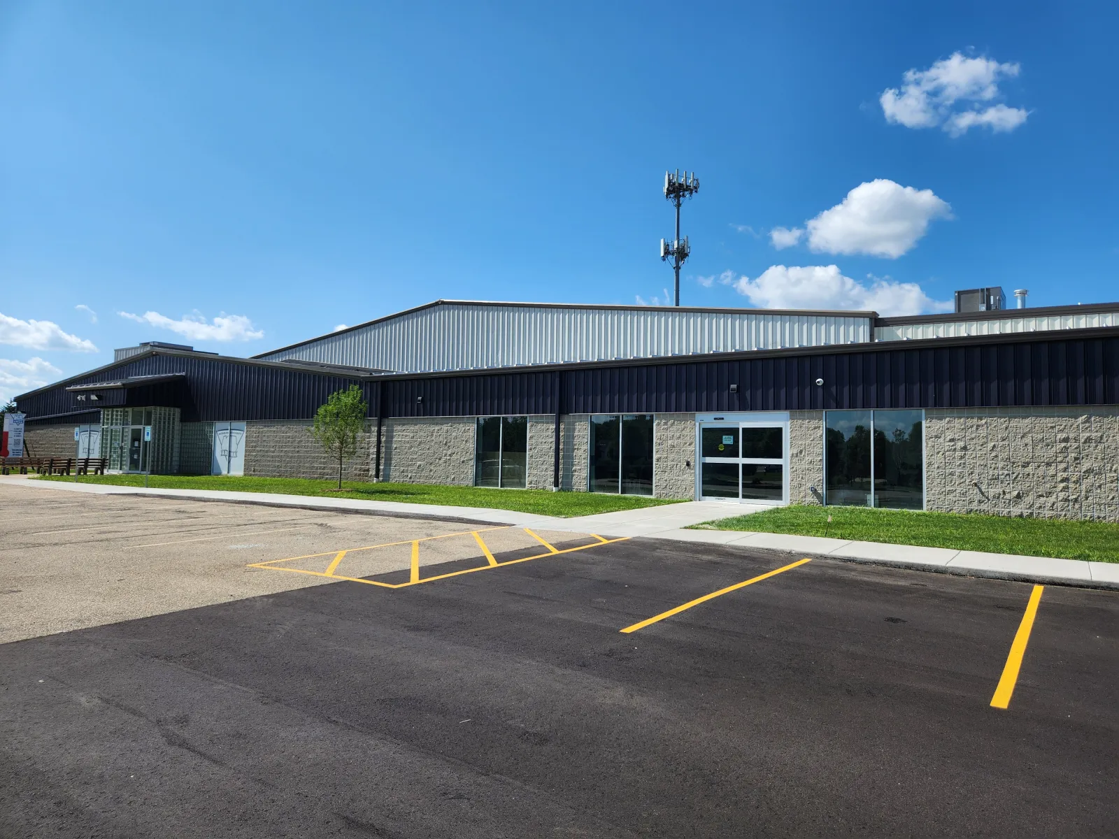 The arena's new stone-and-glass entrance addition and long dark-blue metal hall behind it, seen across a freshly striped parking lot under a blue sky. — McFarland Community Ice Arena