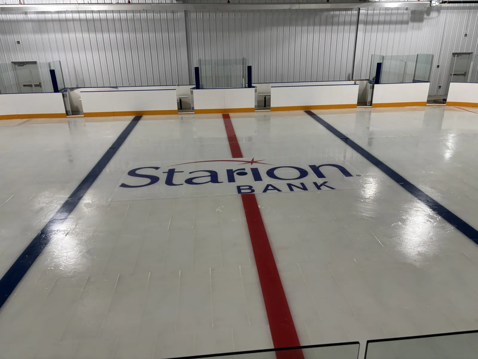 The empty Starion Bank training rink: fresh white ice with the Starion Bank logo at centre, red and blue lines, and white boards with glass along the far side. — McFarland Community Ice Arena