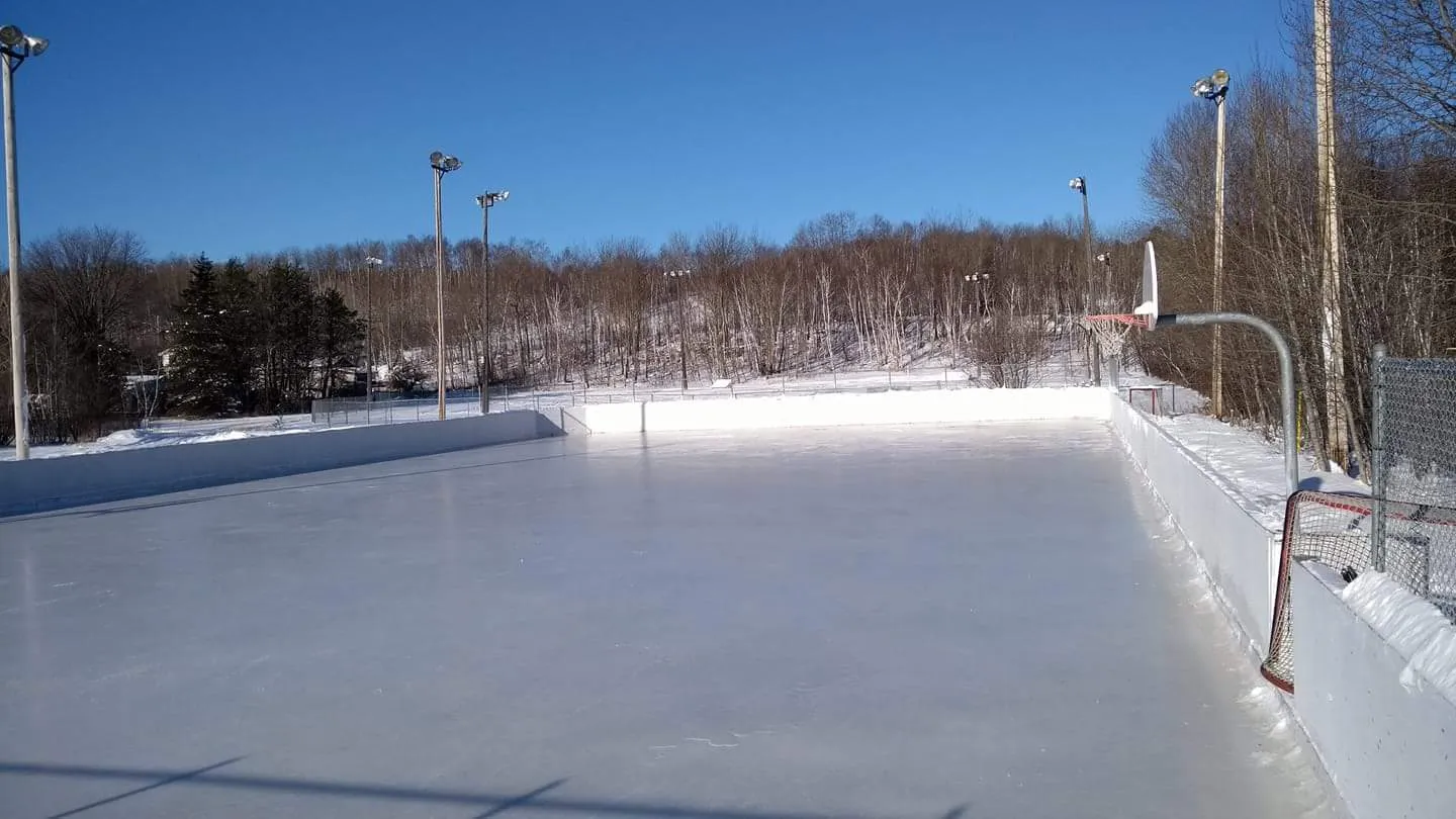 The boarded outdoor rink with a clear ice surface, light poles and a tree line behind it. — McFarlane Lake Playground and Complex Outdoor Rink