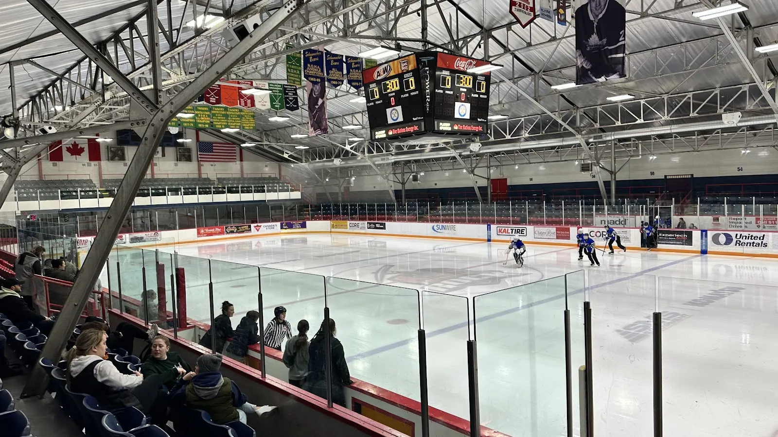 The McIntyre Arena ice seen from the stands, with a few skaters at the far end, the scoreboard above centre ice and flags and banners along the roof. — McIntyre Community Centre (Arena)