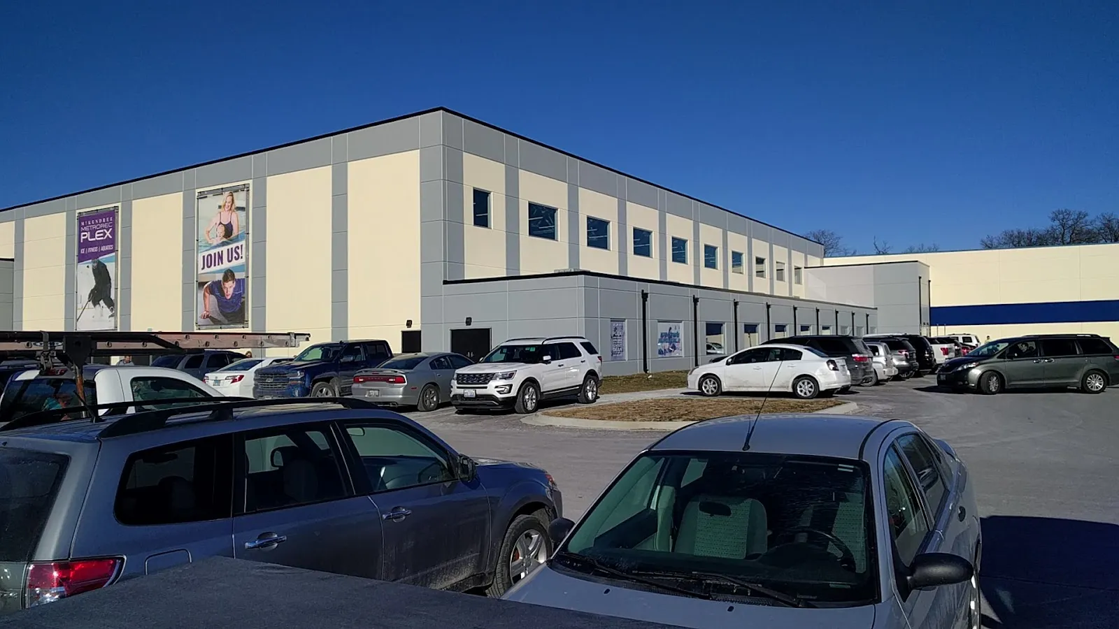The long side of the recreation center building, a pale panelled wall with a row of blue windows and a Plex banner, seen from the parking lot with cars in front. — McKendree Metro Rec Plex
