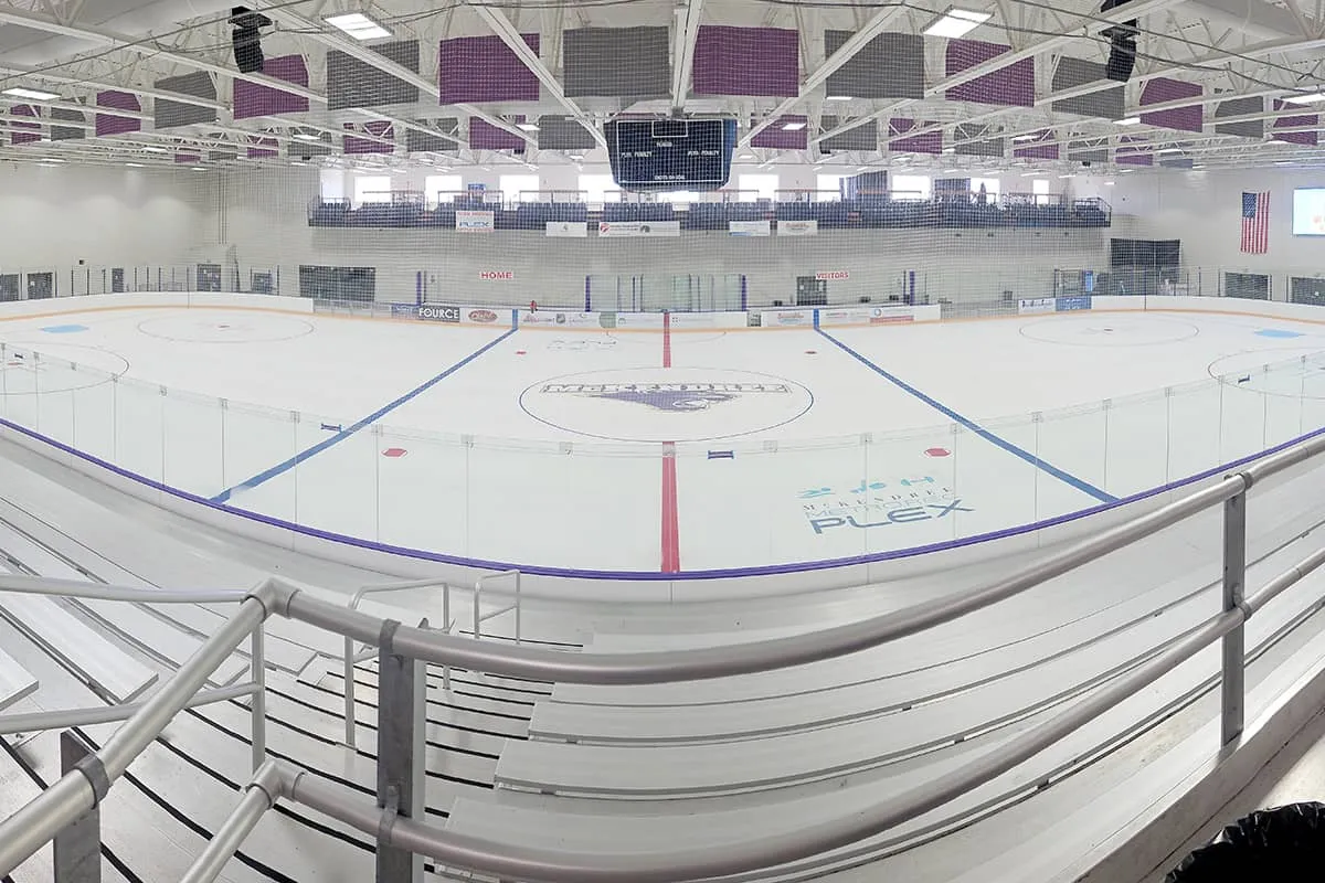 Panoramic view of an empty ice sheet from the bleachers, with a McKendree logo at center ice, the Metro Rec Plex logo inside the near boards, a center-hung scoreboard and blue seats on the far balcony. — McKendree Metro Rec Plex