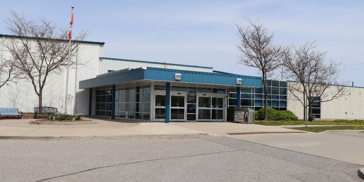 La entrada del McKinney Centre en primavera: un vestíbulo bajo con fachada de vidrio y una marquesina azul frente a los altos muros blancos de la arena, con árboles sin hojas y un asta de bandera. — McKinney Centre