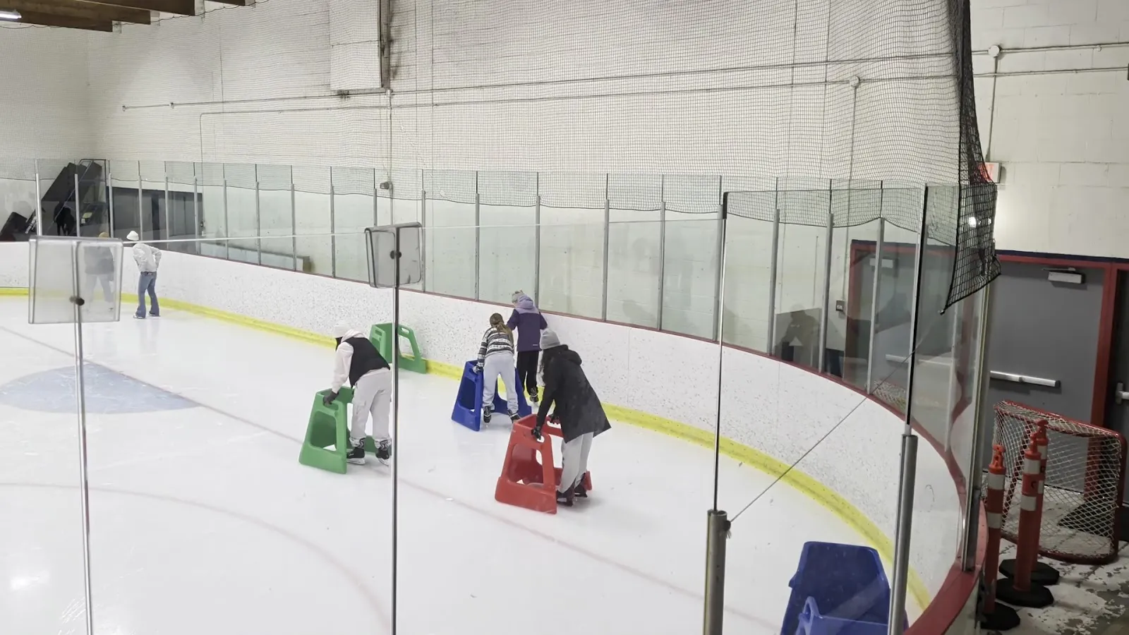 Children skating with plastic skating aids on the ice at McLaren Arena, seen through the glass, with the cone-lined boards and netting beside them. — McLaren Arena