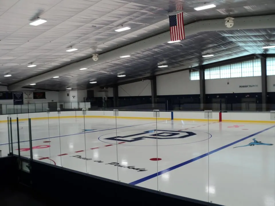 Pista de hielo vacía bajo el techo metálico de la pista, con un gran logotipo de "P" en el centro, las letras "Pride in Peabody", una bandera de Estados Unidos colgada de las armaduras del techo y letreros de los "Peabody Tanners" en la pared del fondo. — James McVann-Louis O'Keefe Memorial Rink