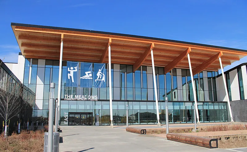Front entrance of The Meadows Community Recreation Centre, a glass-fronted building under a wood-slat canopy roof, with wall signage reading 'THE MEADOWS COMMUNITY RECREATION CENTRE' and an 'EAST' entrance marker, benches out front. — The Meadows Arena