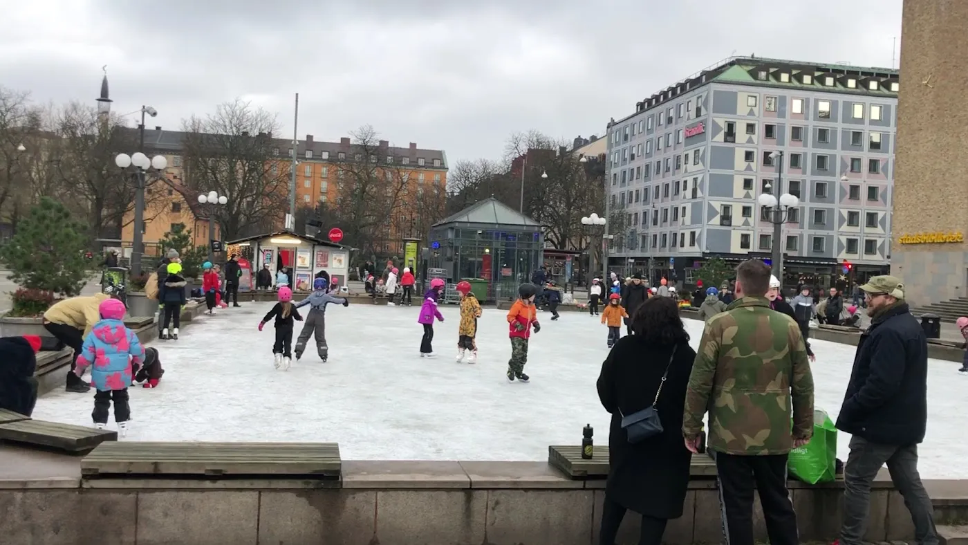 The open ice of Medborgarplatsens isbana at the square seen from the edge, the rink surrounded by a low stone wall with the Södermalm buildings and a pavilion in the background — Medborgarplatsens isbana