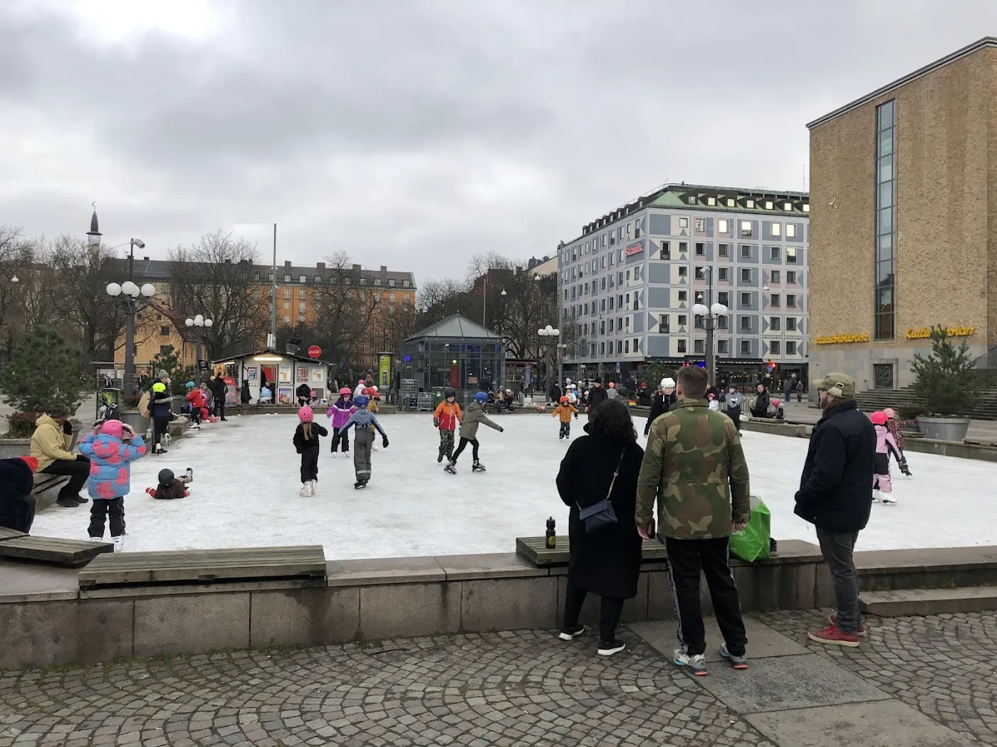 Children and families skating on the artificial ice of Medborgarplatsens isbana in winter, seen over the low stone wall with the square's buildings behind — Medborgarplatsens isbana