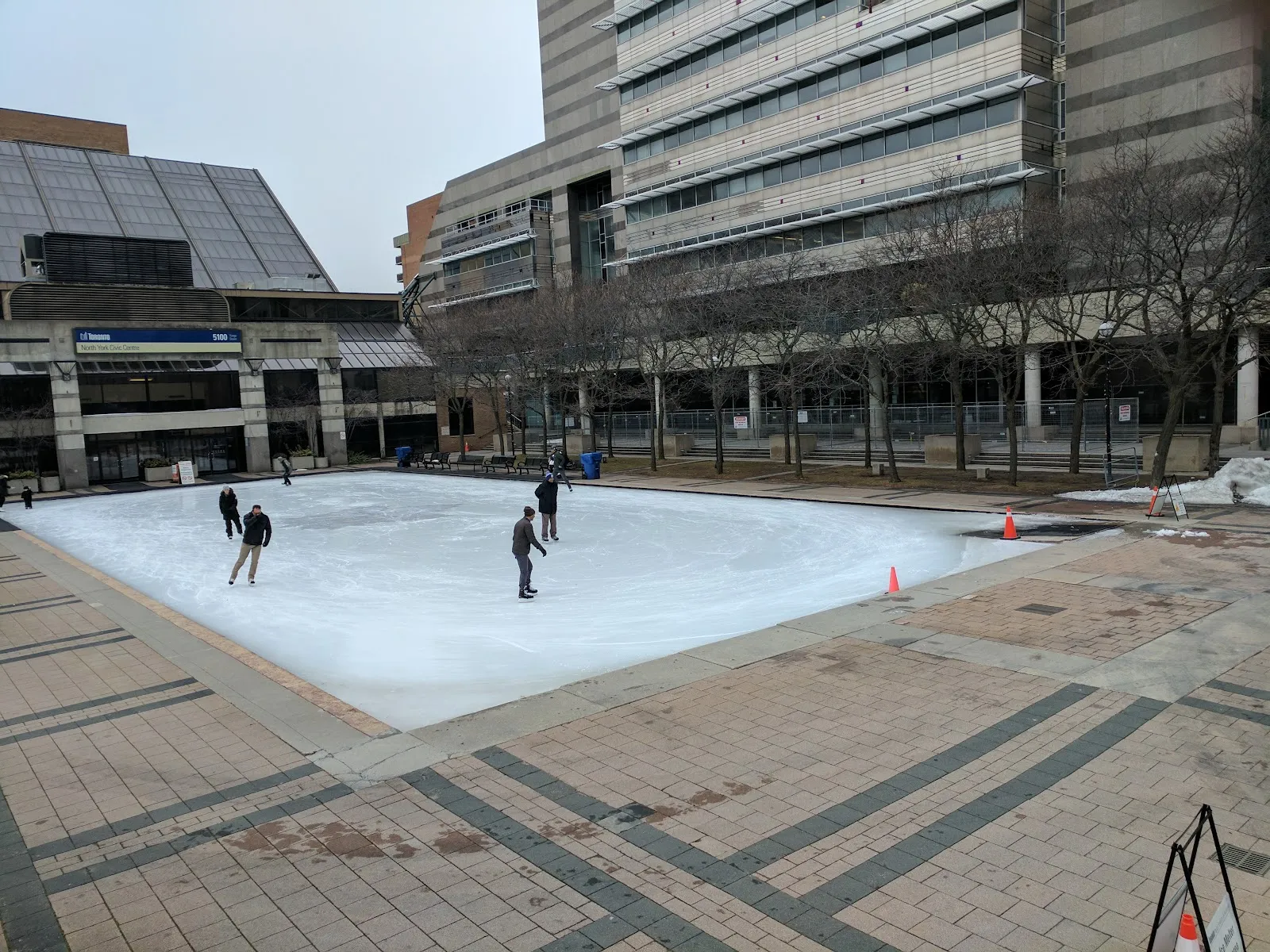 The Mel Lastman Square rink freshly frozen, with a few skaters on the ice, brick paving and the North York civic buildings behind — Mel Lastman Square Outdoor Ice Rink