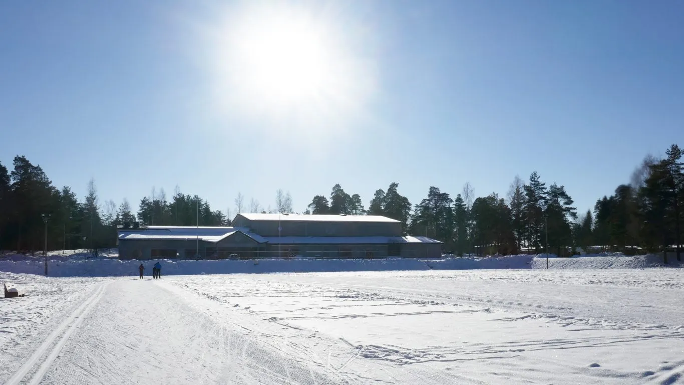 Winter view of the Melassuo sports area with the Perniö sports hall behind a snow-covered field in low sunlight. — Melassuon kaukalo ja luistelukenttä (Salo)