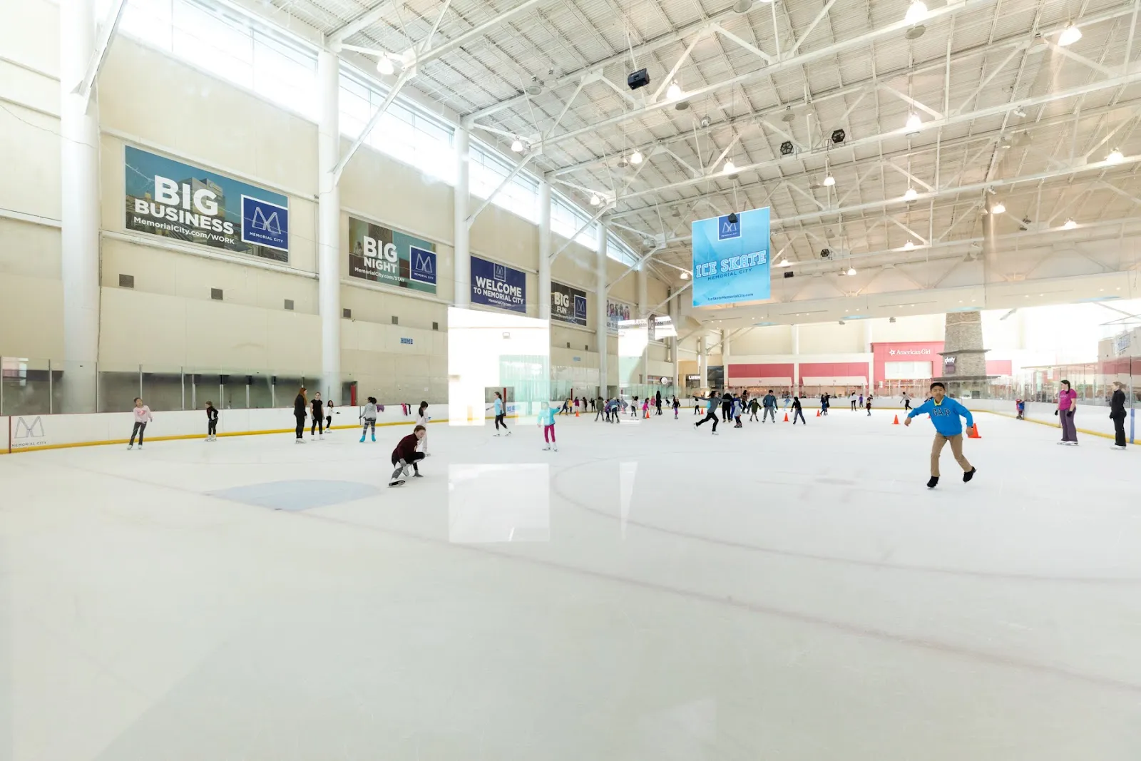 La pista completa de tamaño NHL durante una sesión pública, con patinadores repartidos por el hielo bajo el alto techo blanco y banderines de patrocinadores en la pared del fondo. — Ice Skate Memorial City