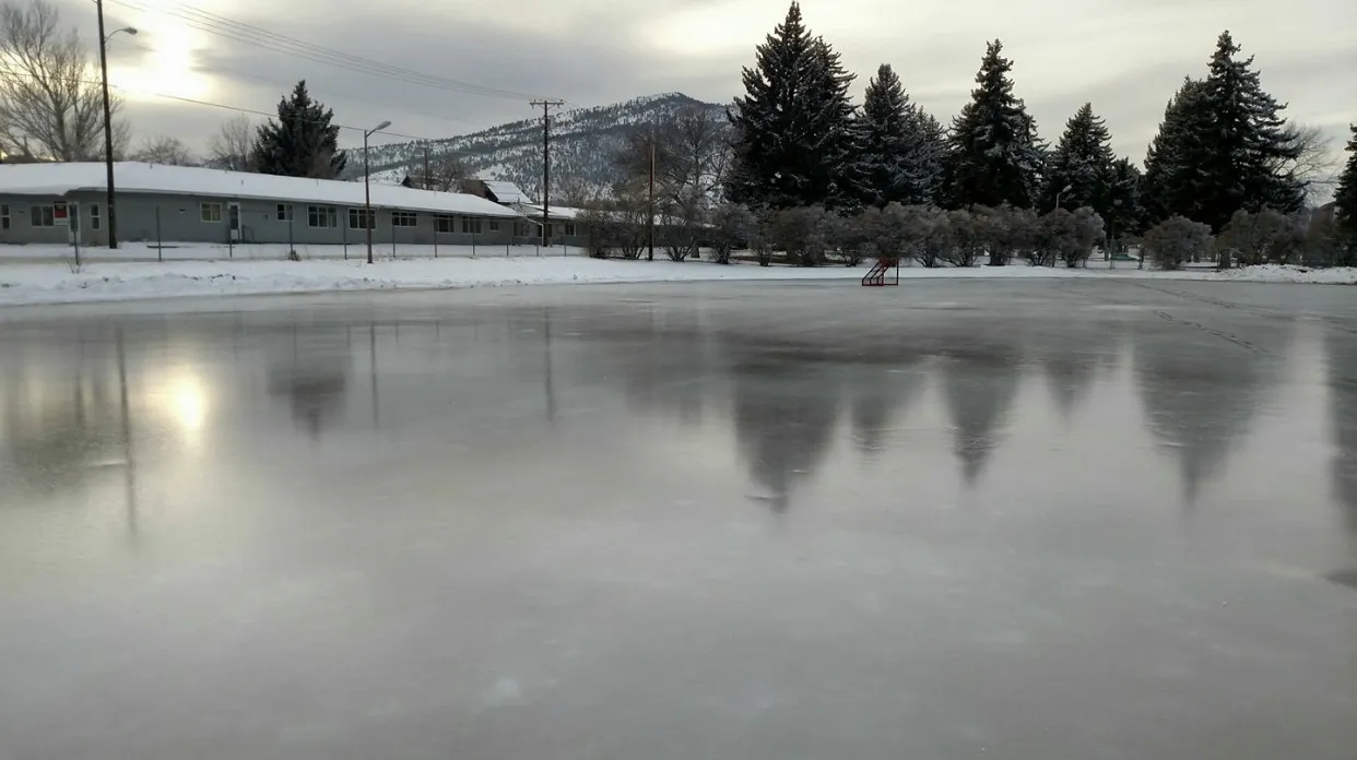 The empty outdoor ice of the Memorial Park Ice Rink seen from the edge in winter, with the park's evergreens and the hills behind. — Memorial Park Ice Rink (Helena)