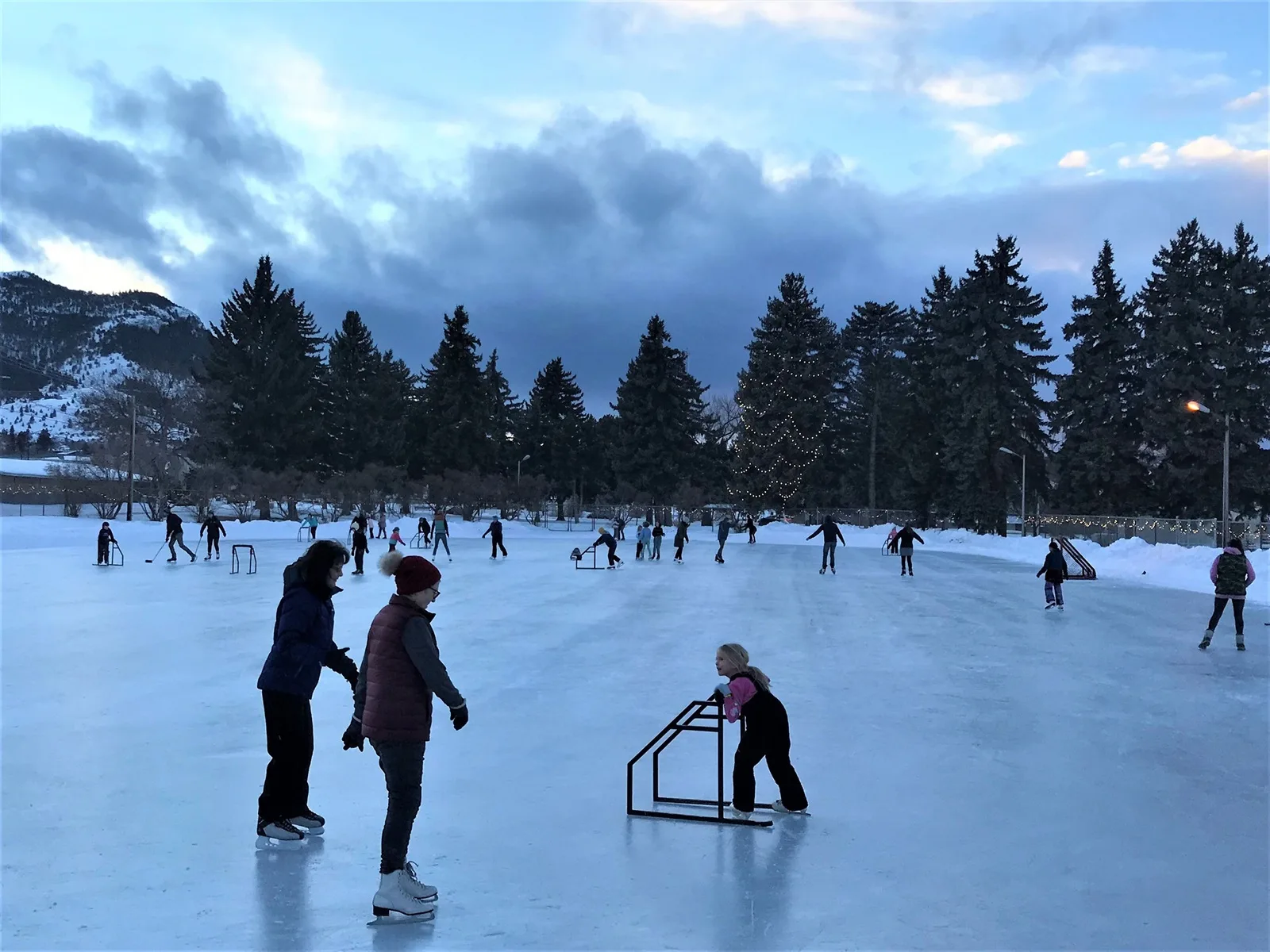 Skaters on the outdoor ice of the Memorial Park Ice Rink at dusk, with skate-aid walkers on the ice, tall evergreens behind and strings of lights along the far side. — Memorial Park Ice Rink (Helena)