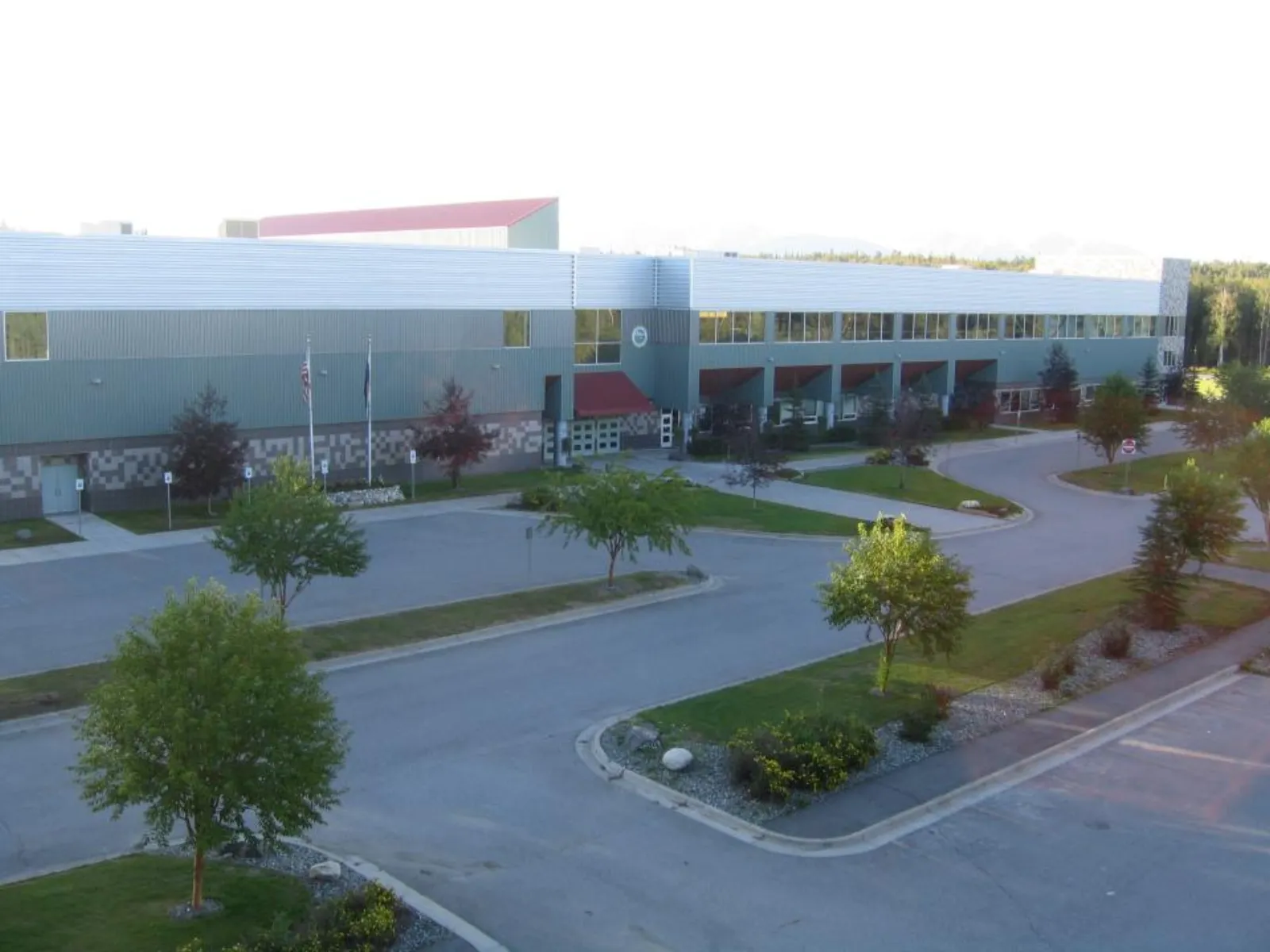 Elevated view of the building's front entrance and parking lot, showing the long metal-panel facade, entrance canopy, flagpoles and landscaped approach. — Curtis D. Menard Memorial Sports Center