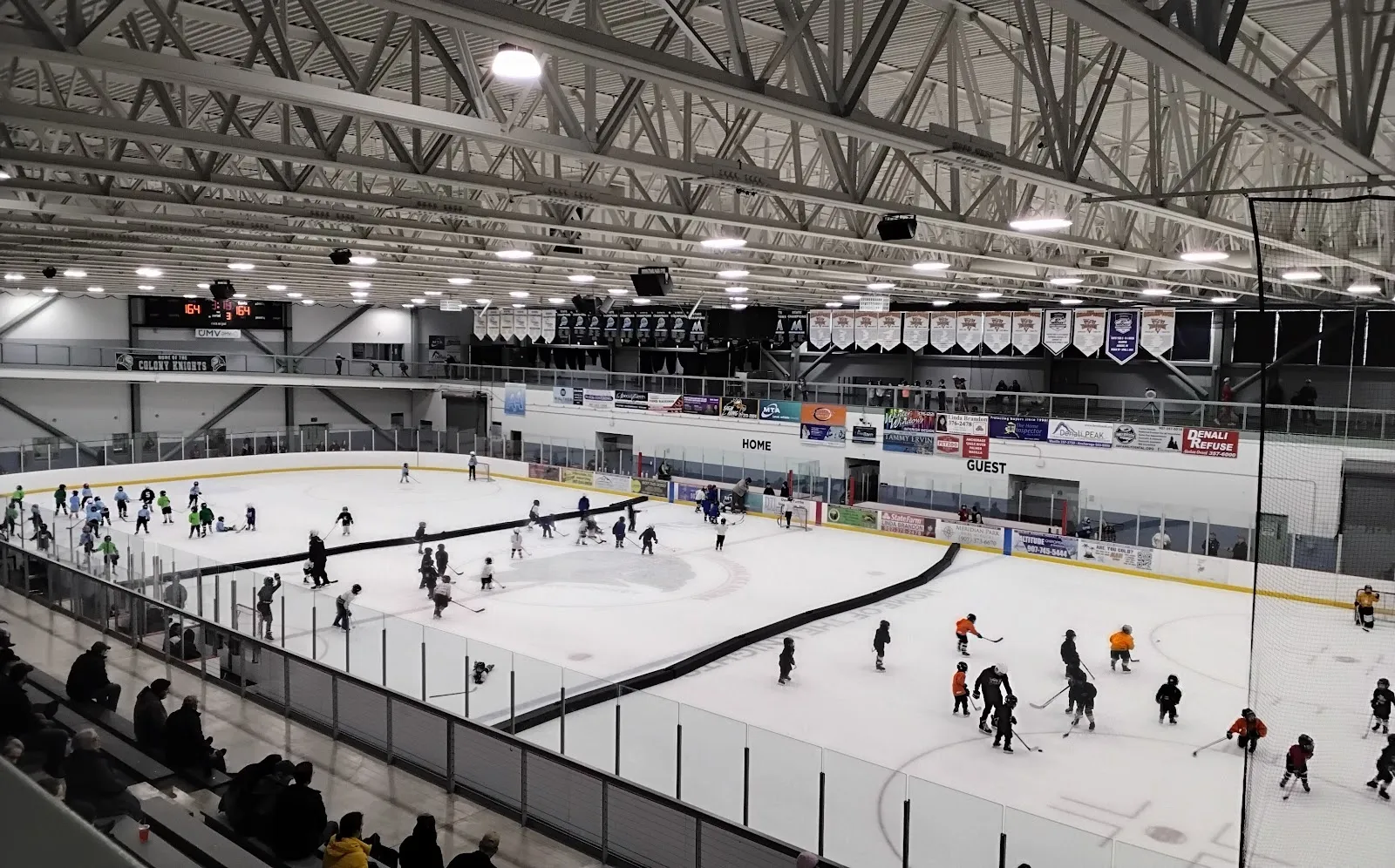 A public skating session seen from the upper rail, skaters spread across a wide sheet in a bright steel-trussed hall with grandstand seating on the near side. — Curtis D. Menard Memorial Sports Center