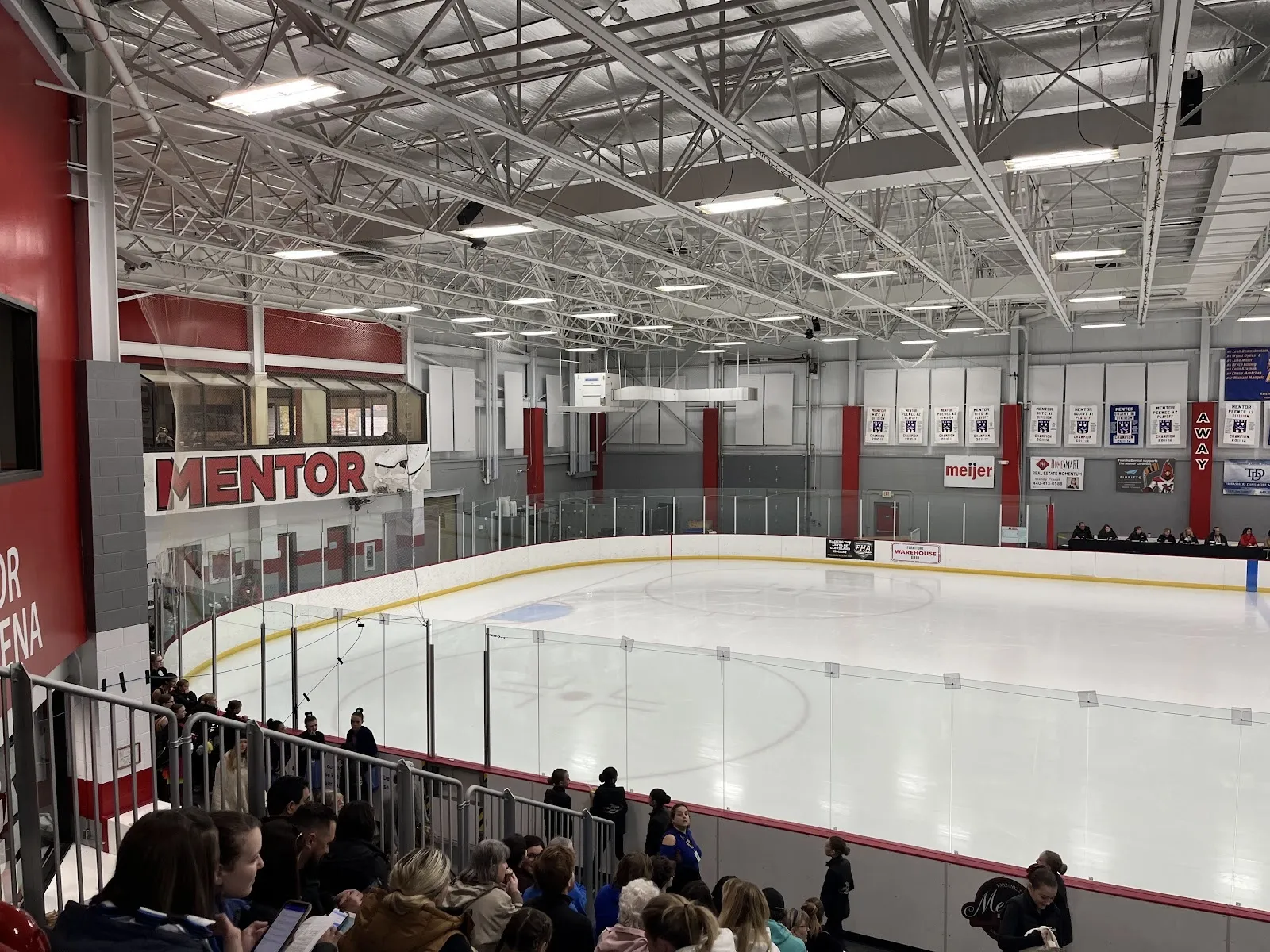 Una vista amplia de la pista desde las gradas, con el hielo vacío curvándose alrededor de las vallas del extremo bajo un techo de armaduras de acero con banderines de equipos en la pared del fondo. — Mentor Civic Ice Arena