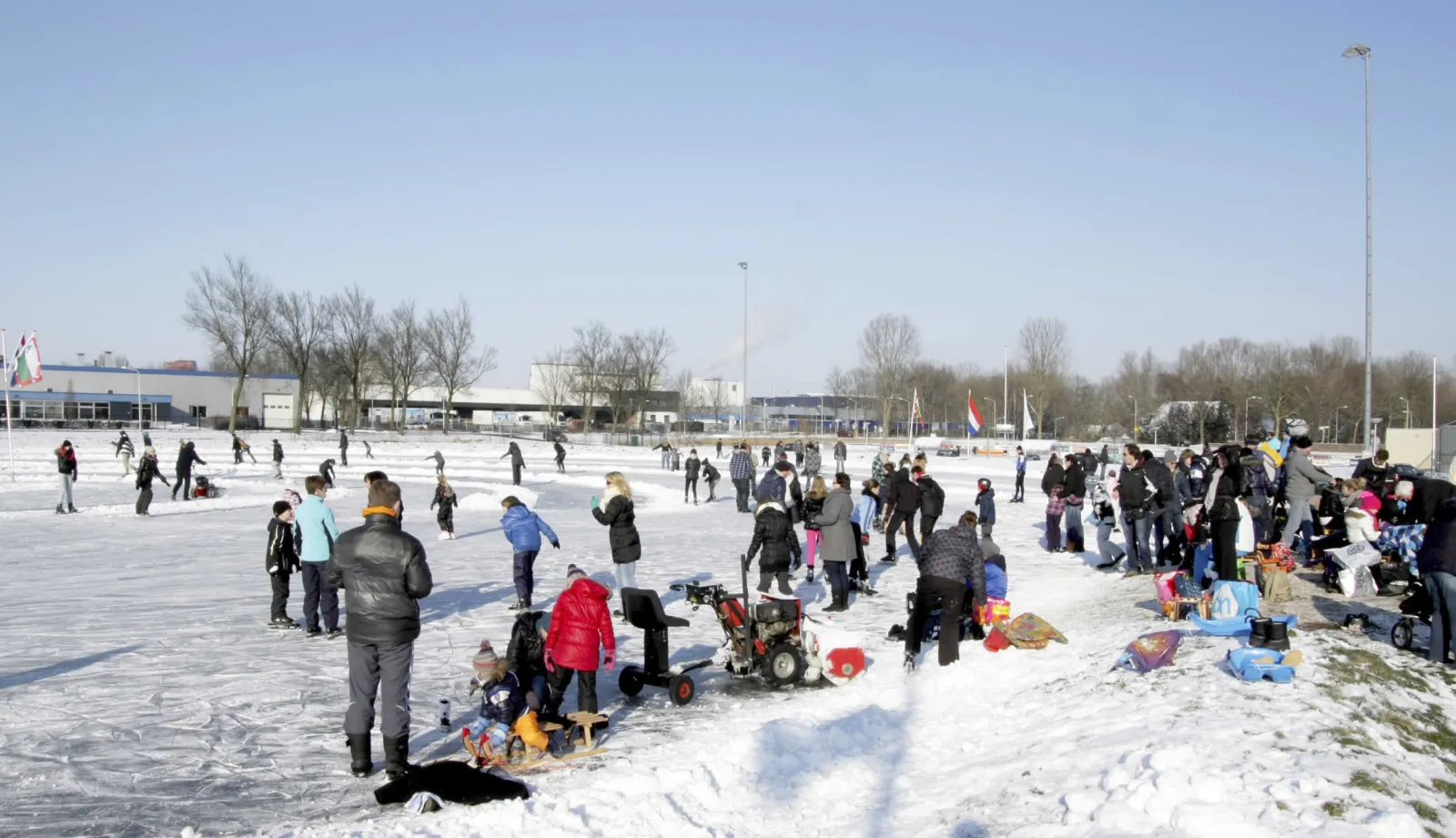 Schaatsers en gezinnen op het besneeuwde natuurijs van Sportpark Koedijkslanden in Meppel op een winterdag, met lichtmasten en de gebouwen van het sportpark erachter. – Meppeler IJsclub