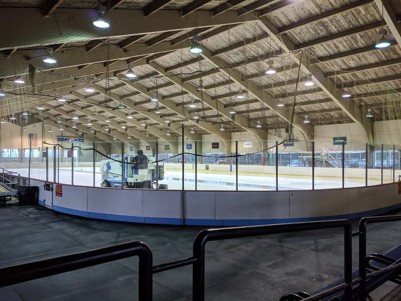 La pista de hielo vista desde detrás de las vallas bajo el amplio techo de madera, con la máquina alisadora sobre el hielo y las gradas de aluminio a la izquierda. — Mercer County Skating Center