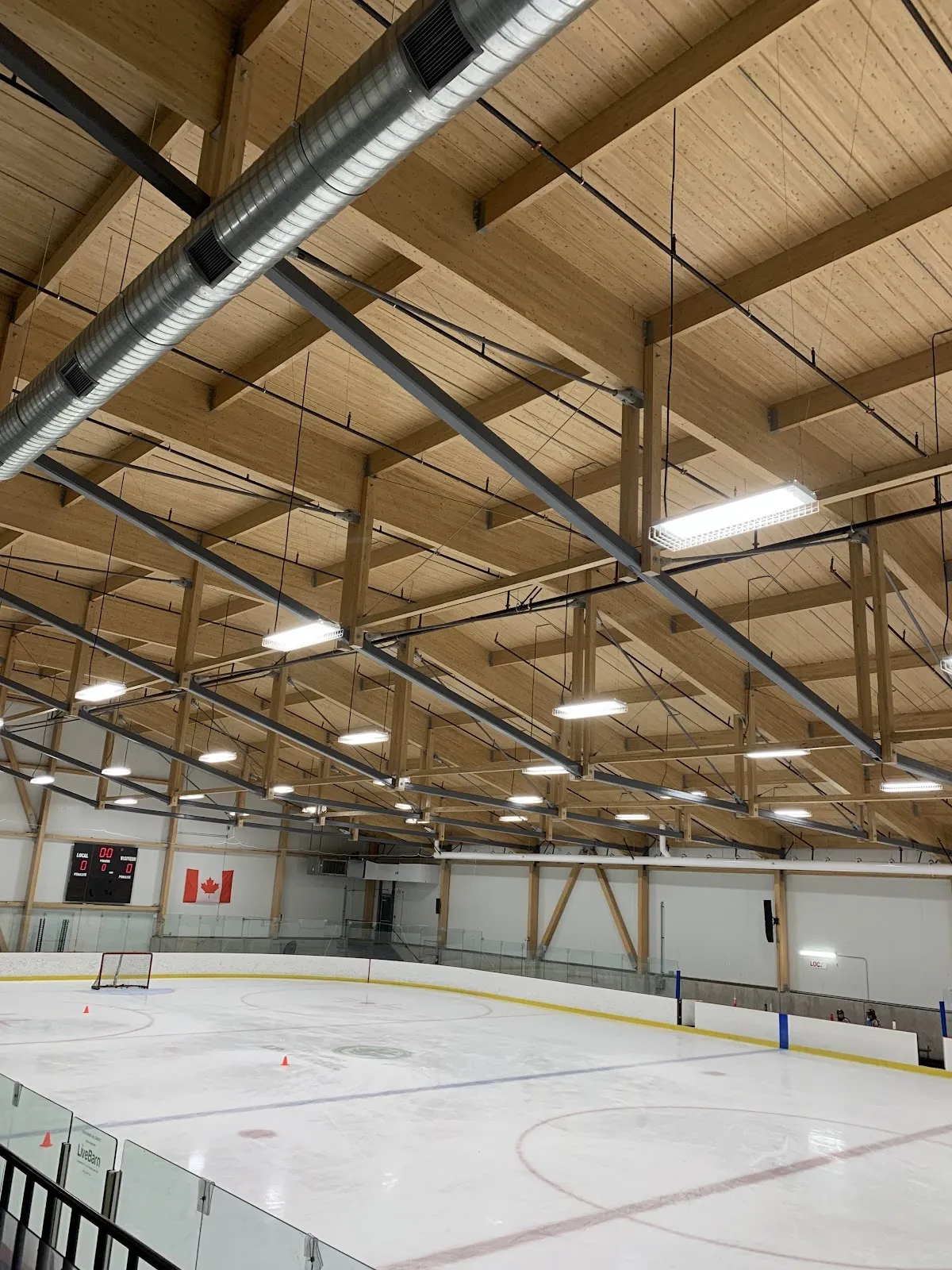 La pista de hielo vacía en el Meredith Centre bajo un techo de madera laminada, con una portería, el marcador y una bandera canadiense a lo largo de las vallas del fondo. — Meredith Centre