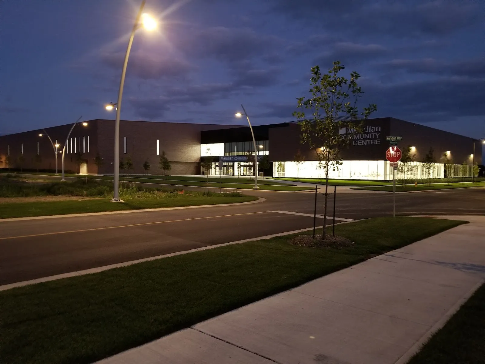 The long, low Meridian Community Centre at dusk, its lit glass entrance and Meridian Community Centre lettering seen across the street from the corner. — Meridian Community Centre